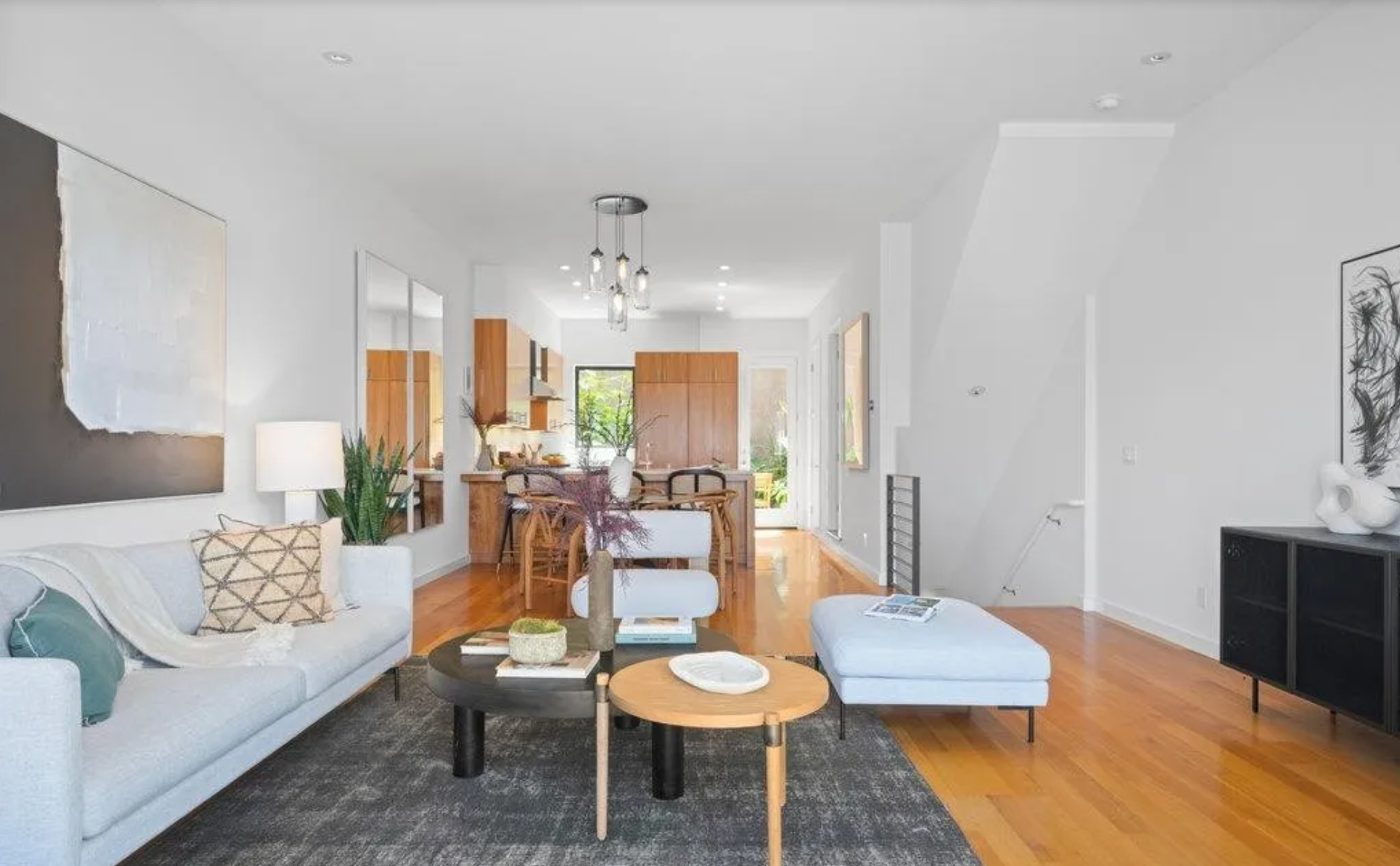 A modern living room features a light-colored sofa, abstract art, potted plants, and nested coffee tables, leading to an open, wooden kitchen with a central island.