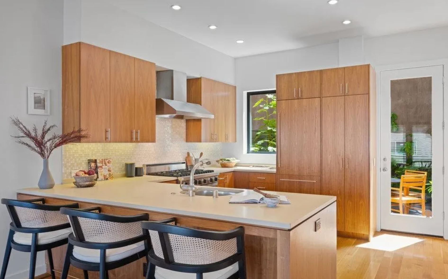 The image depicts a modern kitchen with wooden cabinets, a central island with three chairs, stainless steel appliances, and a door leading to an outdoor space.