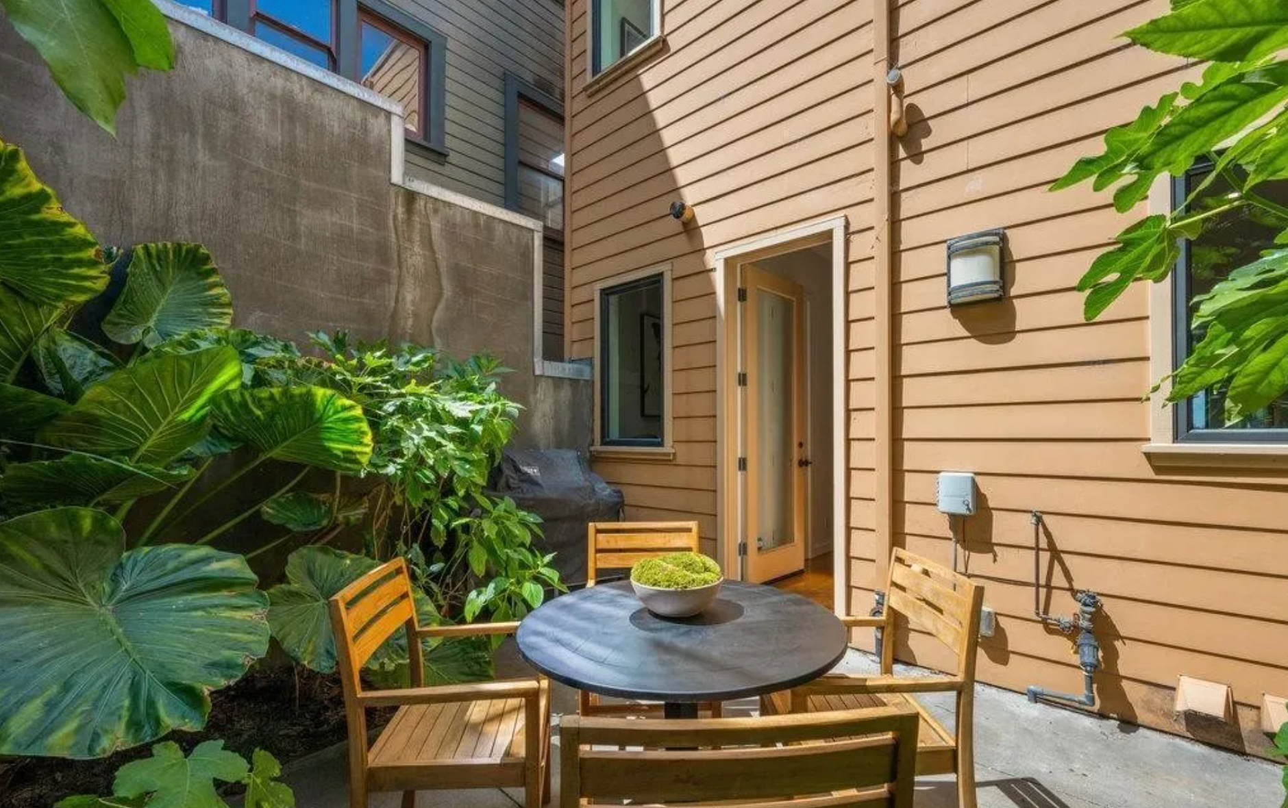 A small backyard patio features a round table with a bowl of plants and four wooden chairs, surrounded by lush green foliage and enclosed by tan wooden siding and a concrete wall.