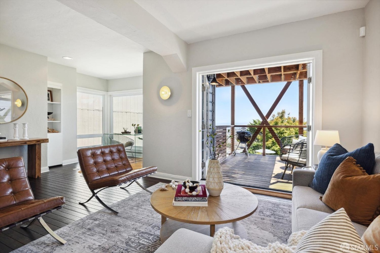 The image shows a modern living room with brown leather chairs, a light-colored sofa, and a round wooden table. Large glass doors open to a balcony with a scenic view.