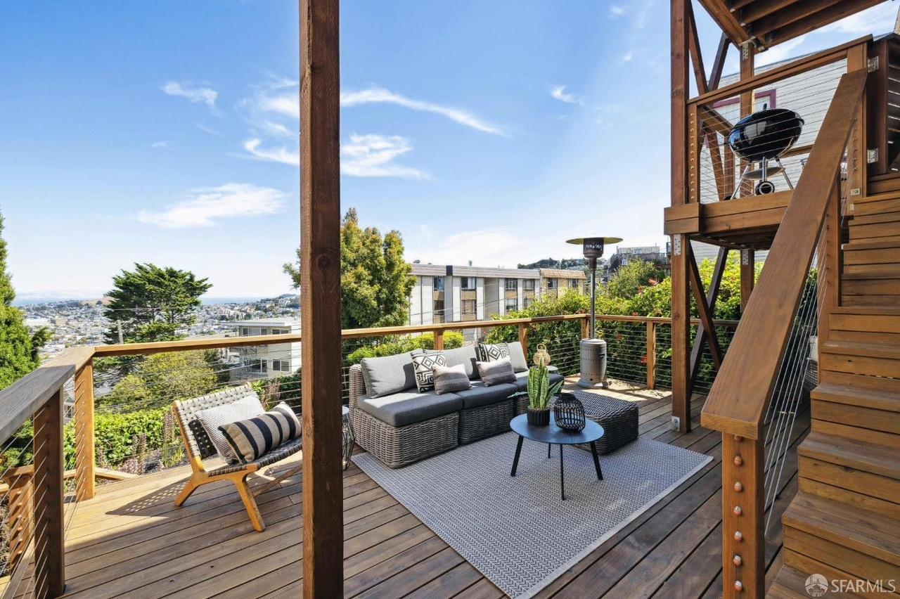 A wooden deck features outdoor furniture, a rug, and a plant, with a railing offering scenic views of the treetops and cityscape under a bright blue sky.