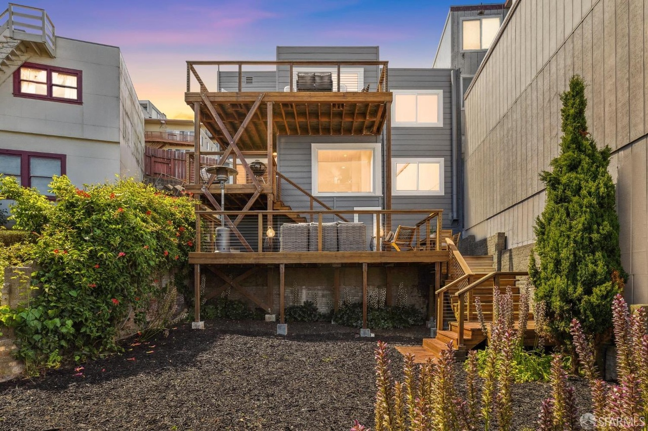 The image shows a three-story house with a wooden terrace and deck at the back, surrounded by neighboring buildings. The yard has a mix of plants and a dark, mulched ground.