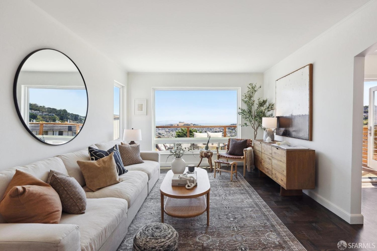 A cozy, sunlit living room with a large window showcasing a scenic view. It features a white couch, wooden coffee table, circular mirror, and a sideboard with decor.