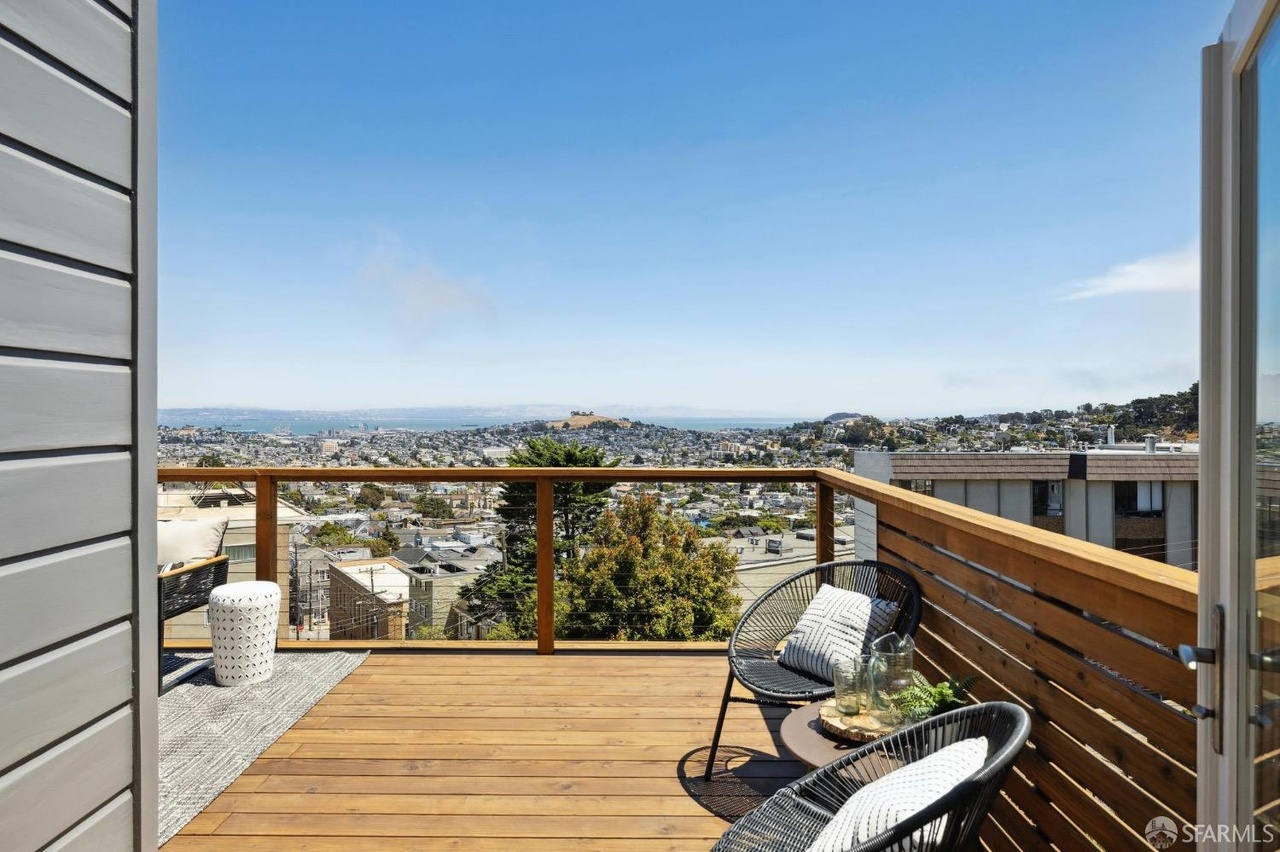 The image shows a wooden balcony with a black metal railing, offering a panoramic view of a cityscape. There are two black chairs with cushions and a small table.