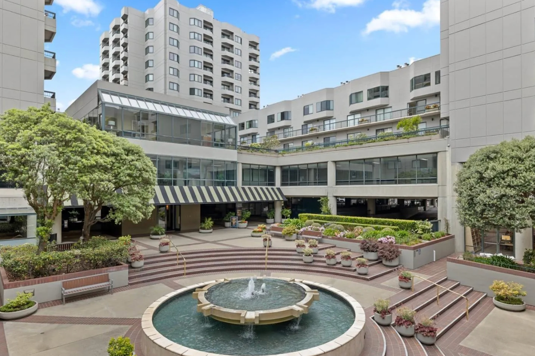 The image shows a courtyard surrounded by modern, high-rise buildings, featuring a central fountain, a staircase, lush greenery, and various planters with flowers.