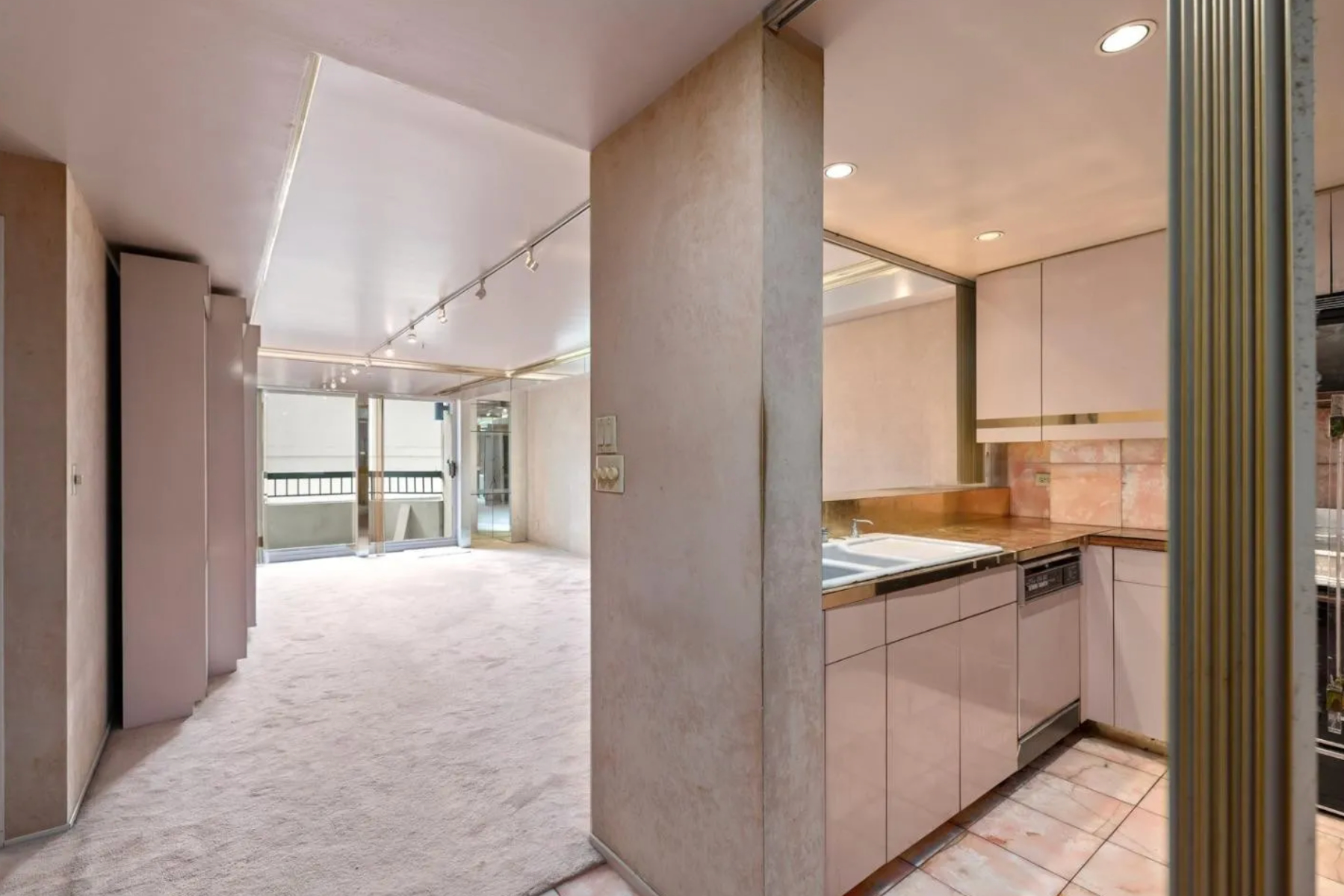 The image shows a narrow, beige-carpeted living room with large glass doors leading to a balcony, adjacent to a compact, tiled kitchen with light-colored cabinets.