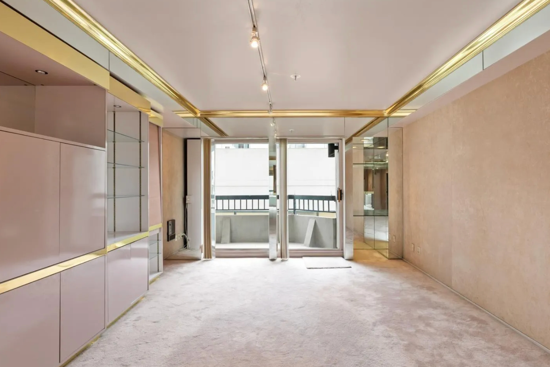 The image shows a modern living room with beige carpeting, gold-accented ceiling, built-in cabinets, mirrored wall panels, and a sliding glass door leading to a small balcony.