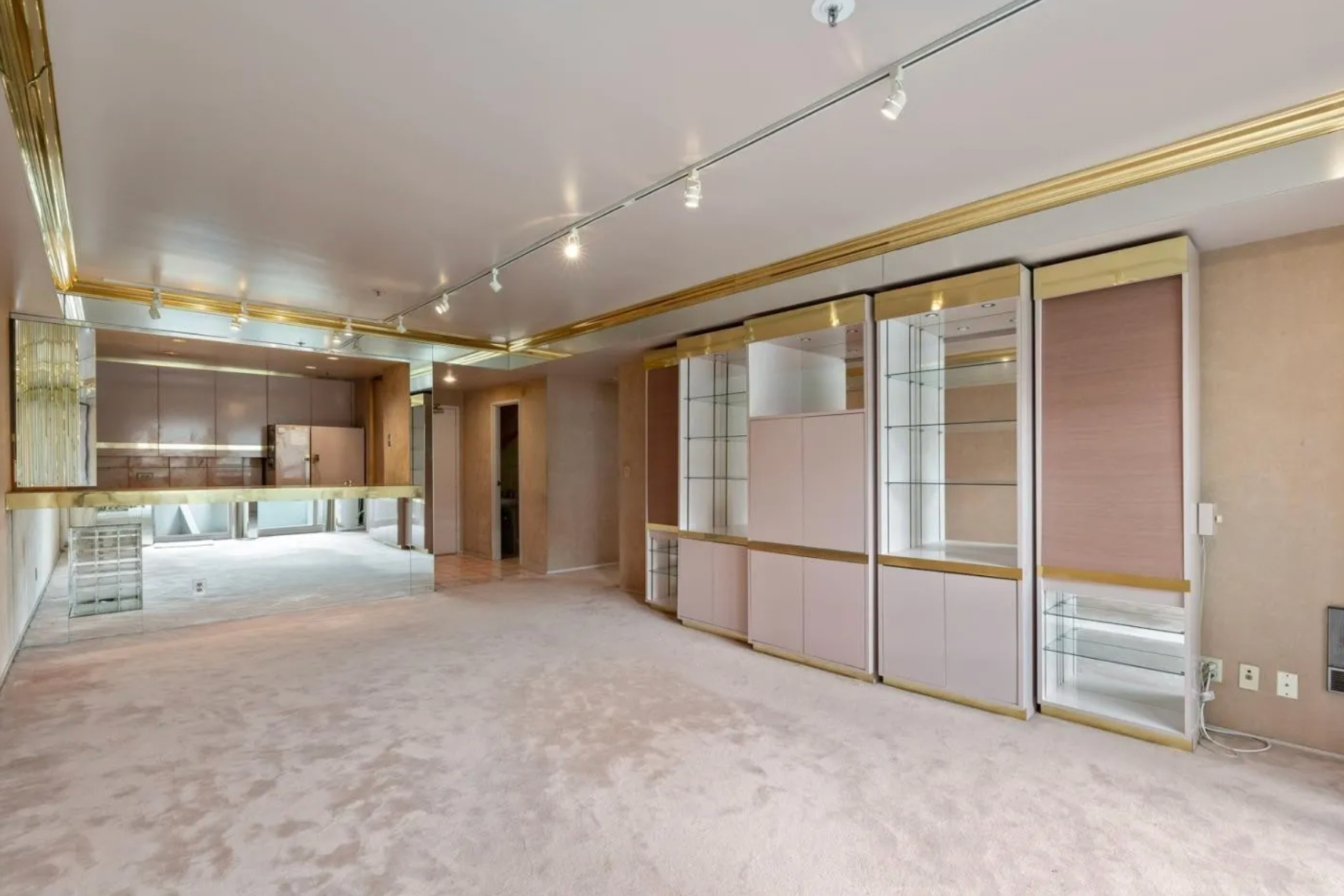 The room has beige carpet, gold accents, built-in glass cabinets with lights, and a mirrored wall section. A partial view of the kitchen and dining area is visible.