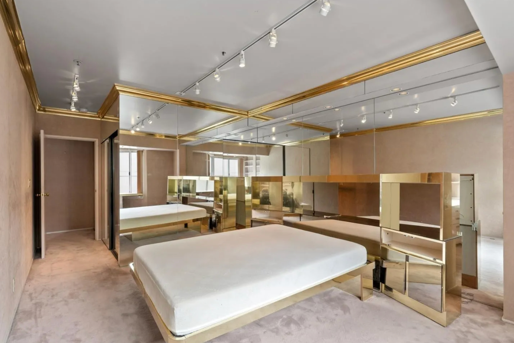 The image depicts a bedroom with beige walls, mirrored closet doors, a low white bed, gold trim, and abundant overhead lighting with track lights.