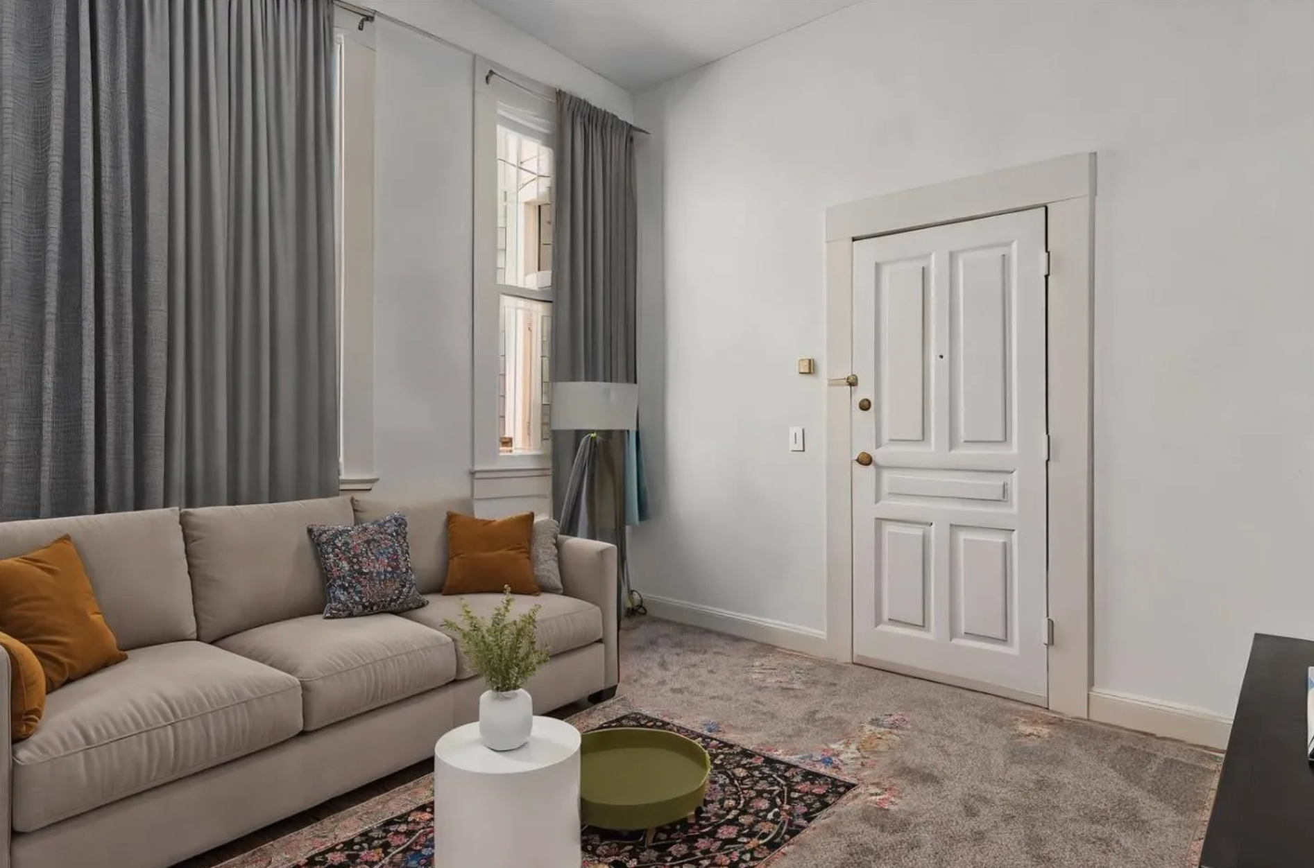 This image shows a living room with a beige sofa adorned with colorful pillows, a small white side table, and a potted plant. There are long gray curtains and a white door.