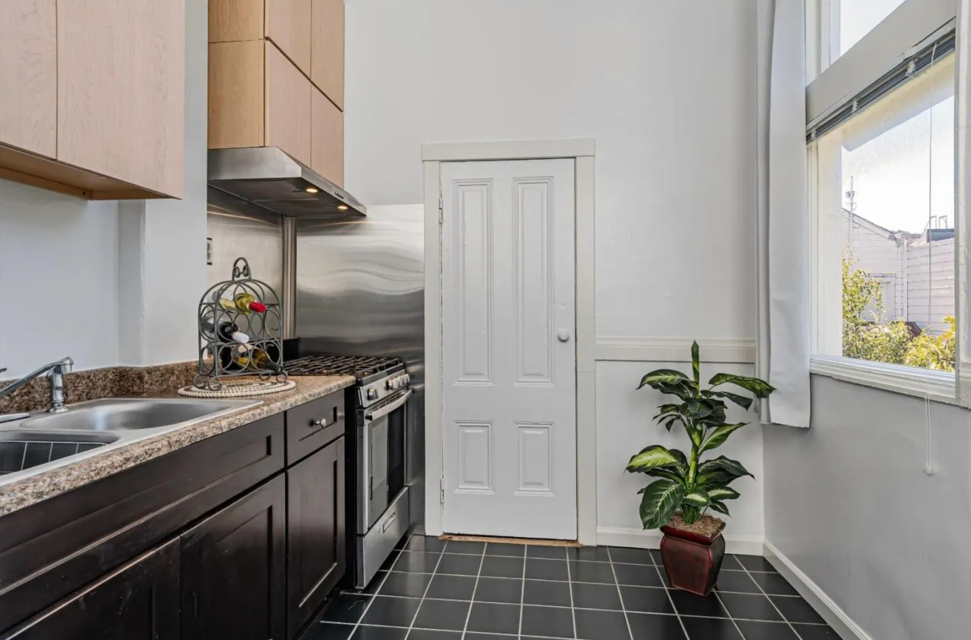 The image shows a modern kitchen with granite countertops, dark cabinets, and a stainless steel stove. A small potted plant sits under a large window, and a wine rack is on the counter.