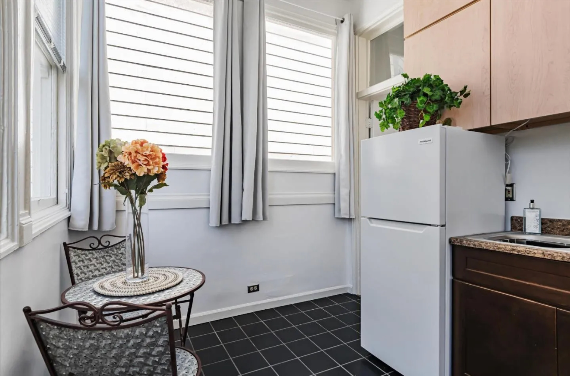 A small kitchen has a white fridge, wooden cabinets, a plant on top, a round table with a floral vase, metal chairs, and large windows with white curtains.