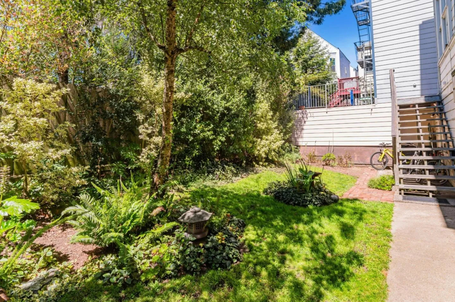 The image shows a small yard with lush greenery, trees, a small path, and a lantern. There is a bike leaning against a wooden staircase, leading up to a two-story building.