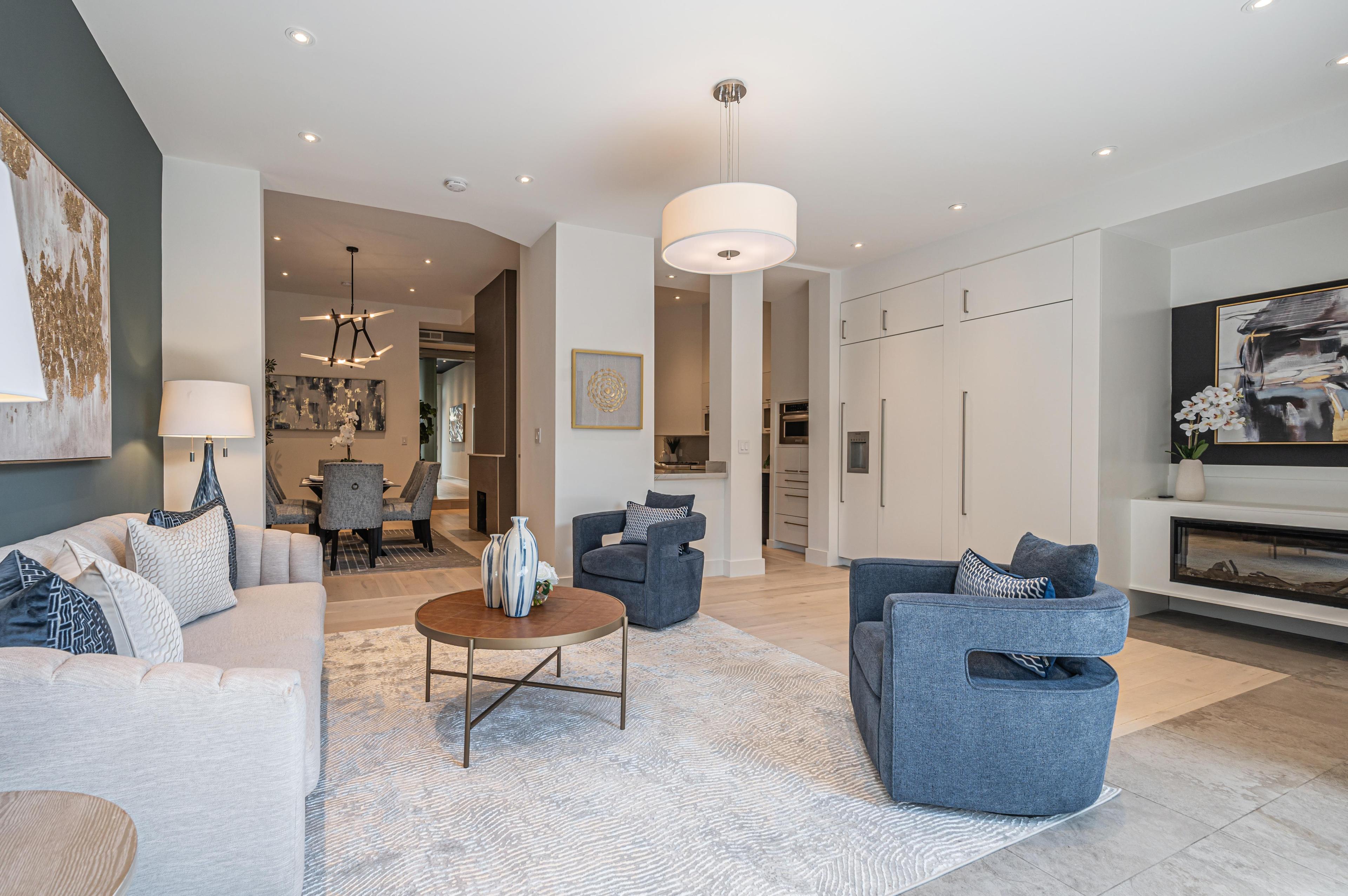 The image shows a modern living room with a grey sofa, two blue armchairs, a round wooden coffee table, a rug, and a dining area in the background.