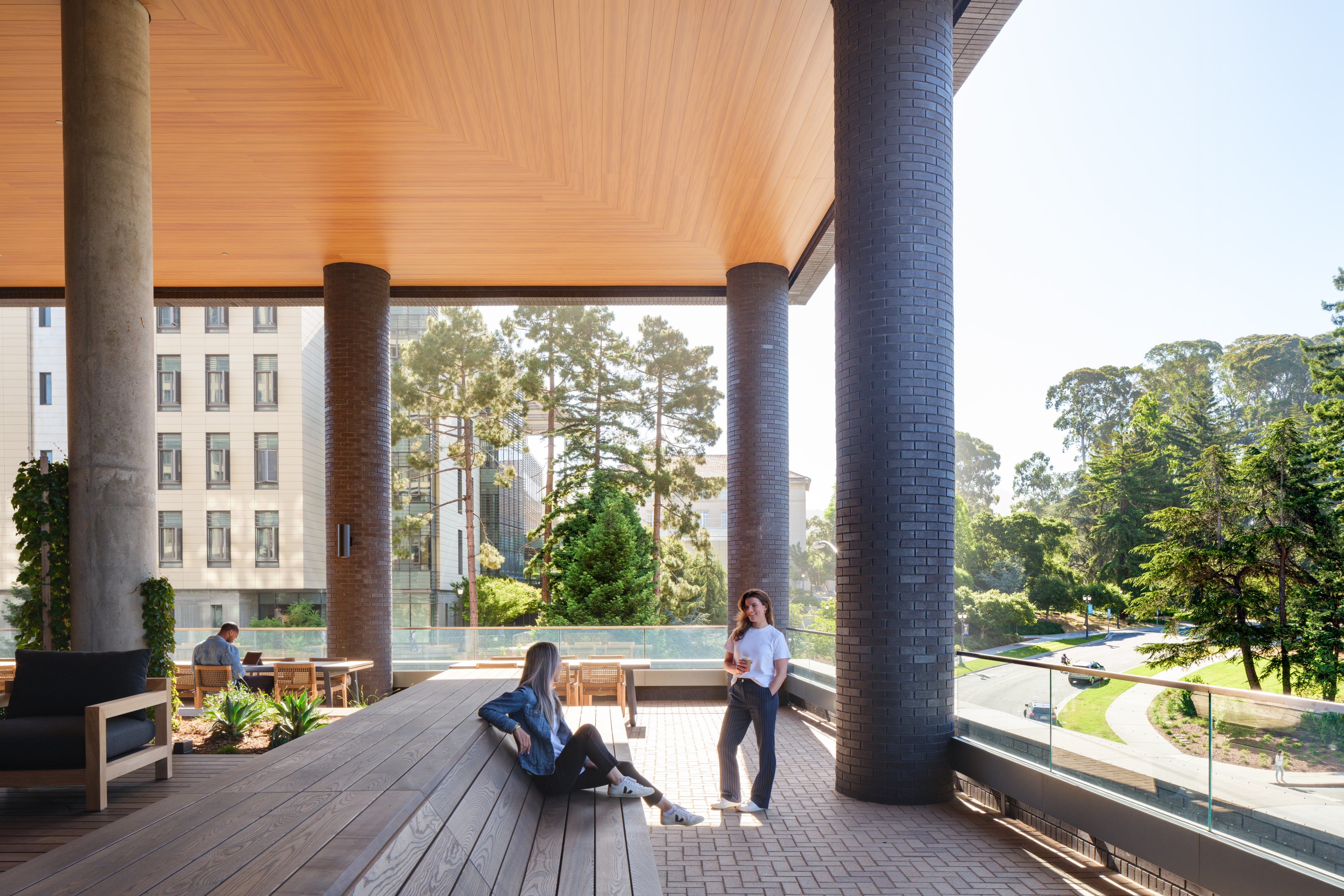 Three people are on a stylish, covered outdoor terrace with wooden seating and large brick pillars, surrounded by a lush, green landscape and modern buildings.