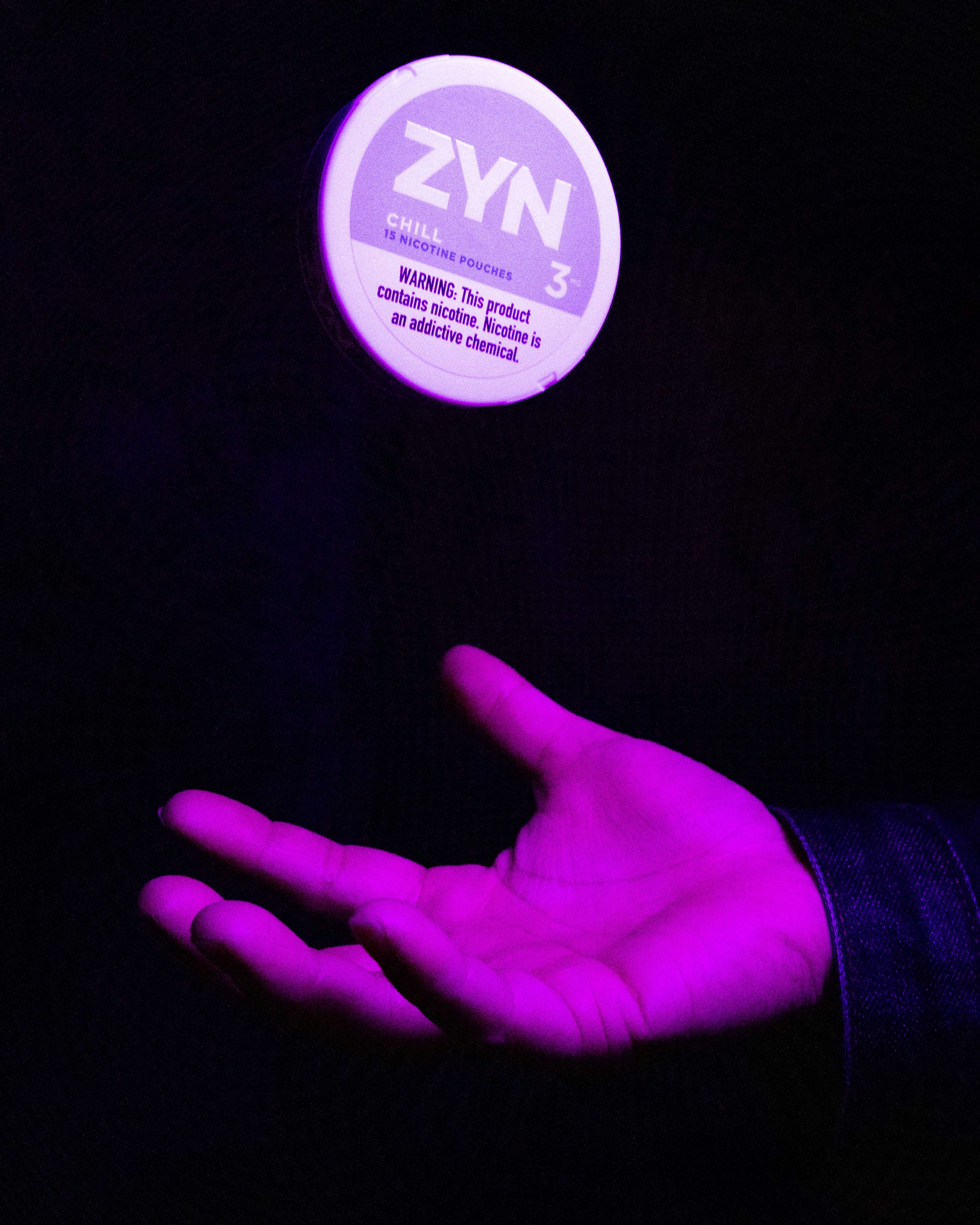 A purple-lit hand appears to be tossing or catching a small round container labeled &quot;ZYN 3,&quot; containing nicotine pouches, against a dark background.