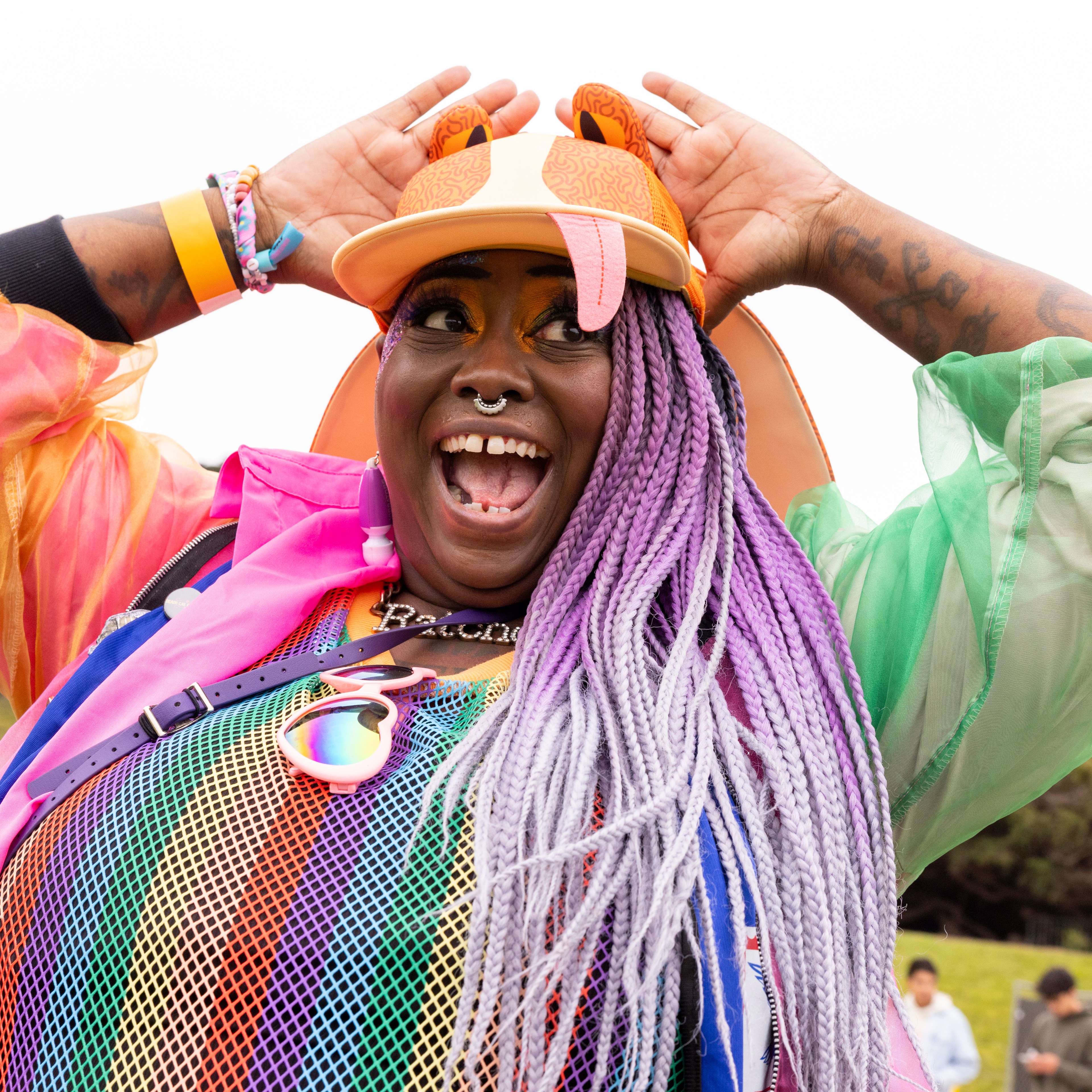 A joyful person with colorful braided hair and a vibrant rainbow outfit poses excitedly with hands on their hat, surrounded by an outdoor event setting.