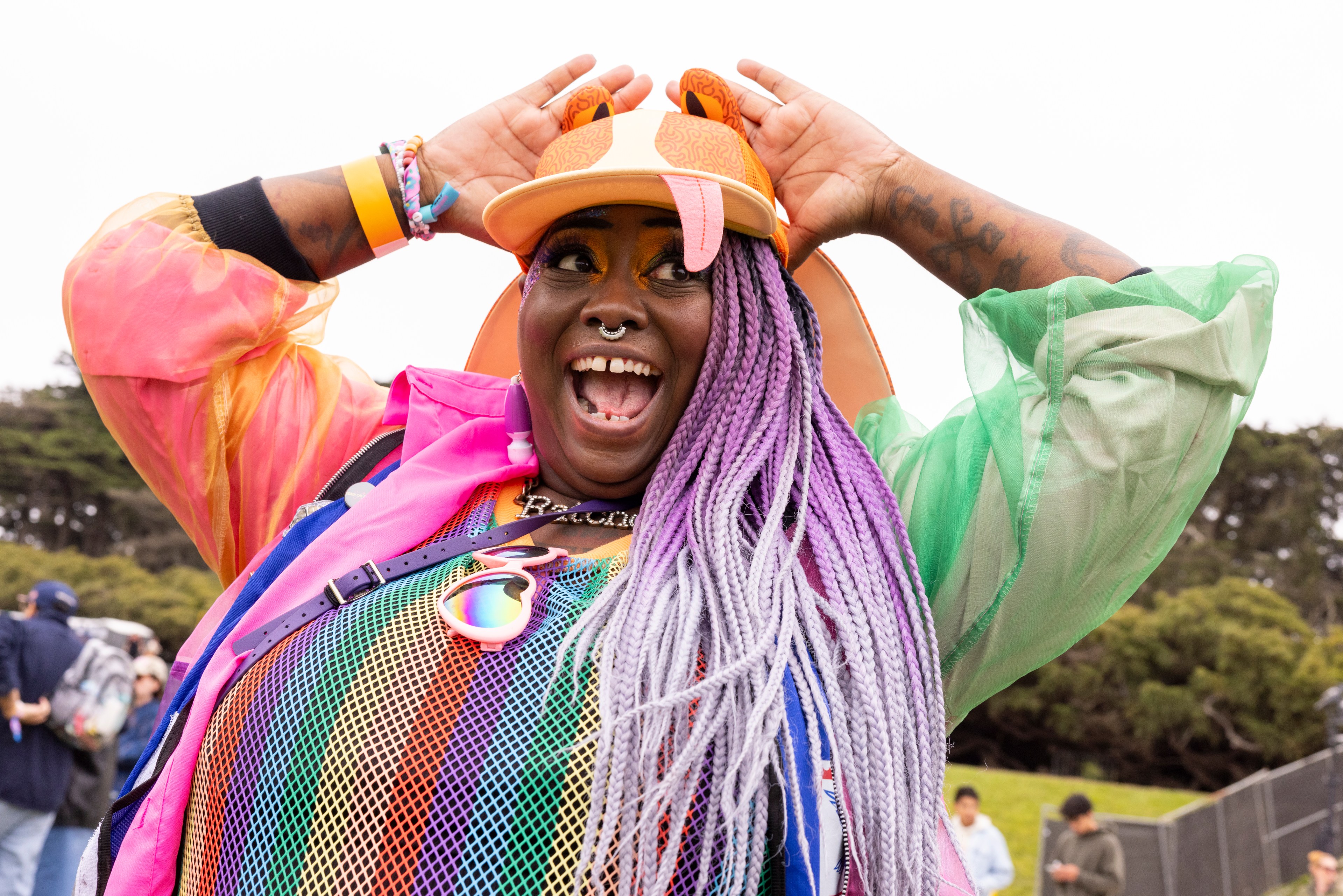 A joyful person with colorful braided hair and a vibrant rainbow outfit poses excitedly with hands on their hat, surrounded by an outdoor event setting.