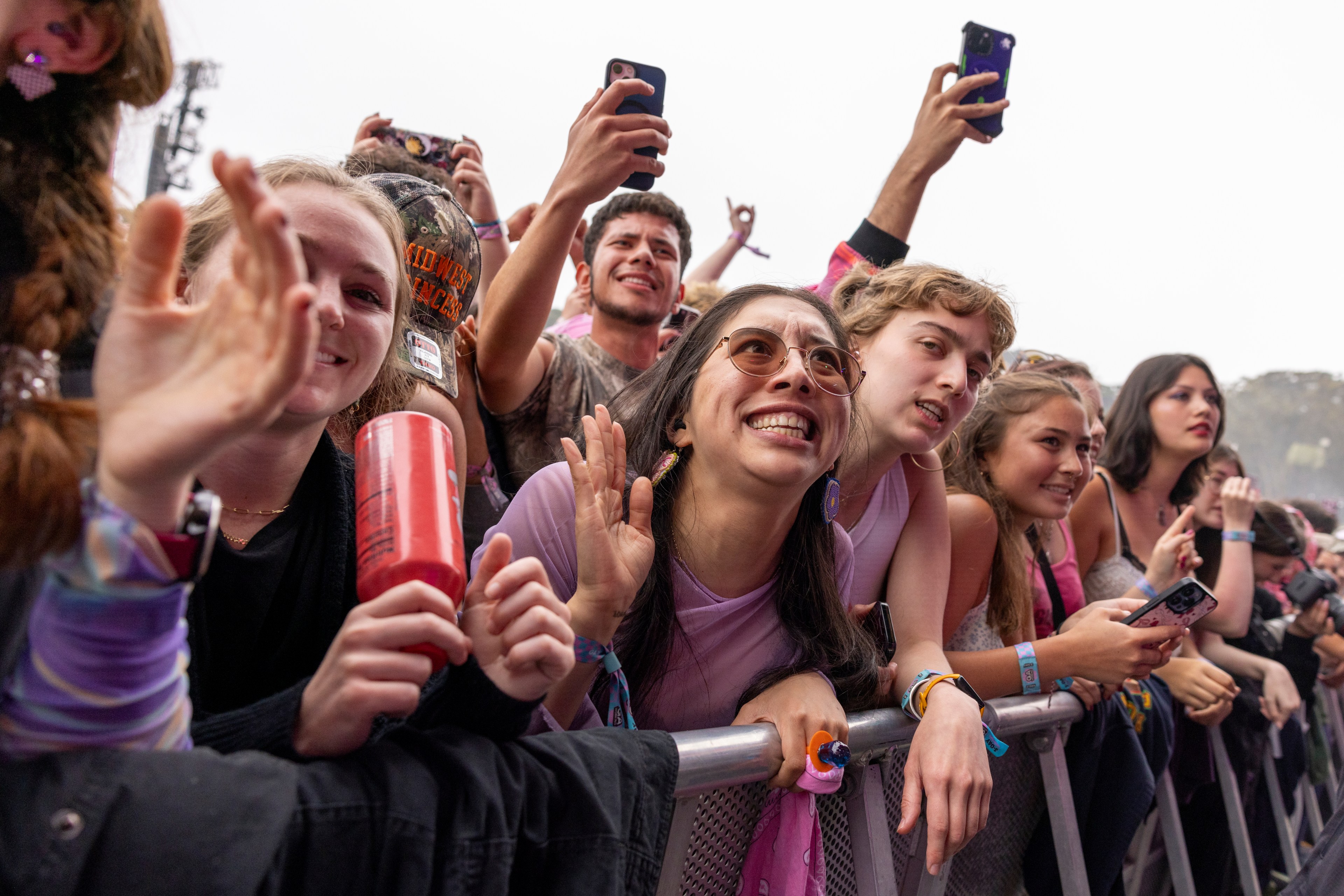 A group of excited young people at a concert stands behind a metal barrier, waving and taking photos with their phones, smiling and cheering energetically.