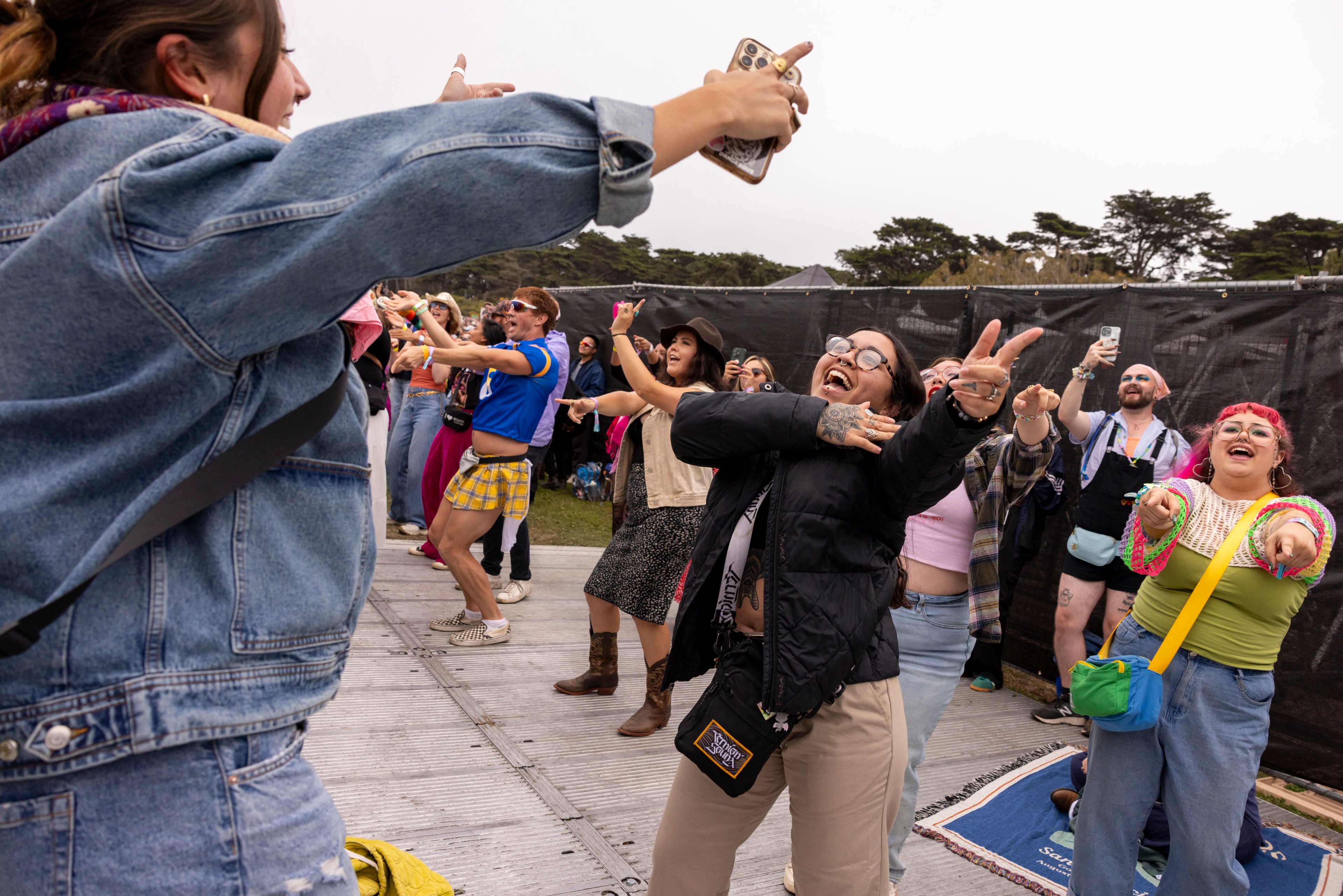 A group of people is excitedly dancing and cheering at an outdoor event, with one person taking a photo. They all exude joy and enthusiasm in various colorful outfits.