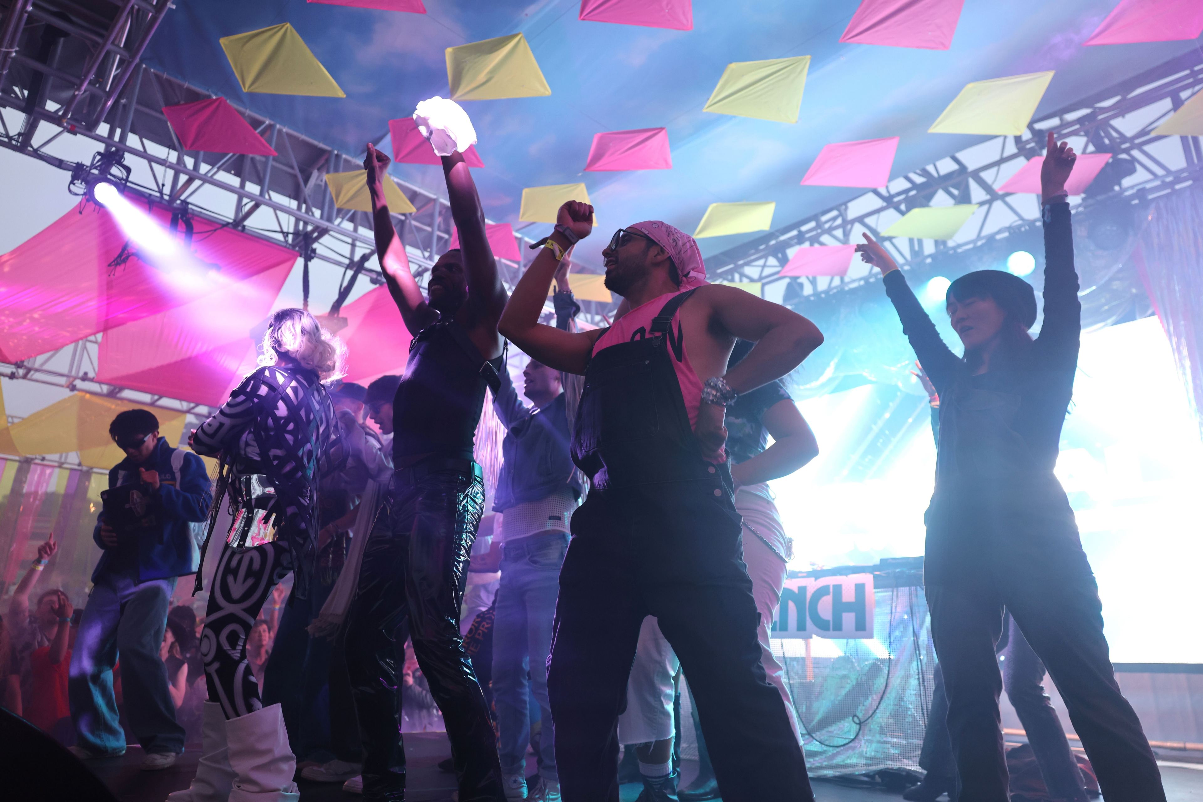 A group of people are dancing energetically on a decorated stage with colorful flags hanging above, illuminated by vibrant lights.