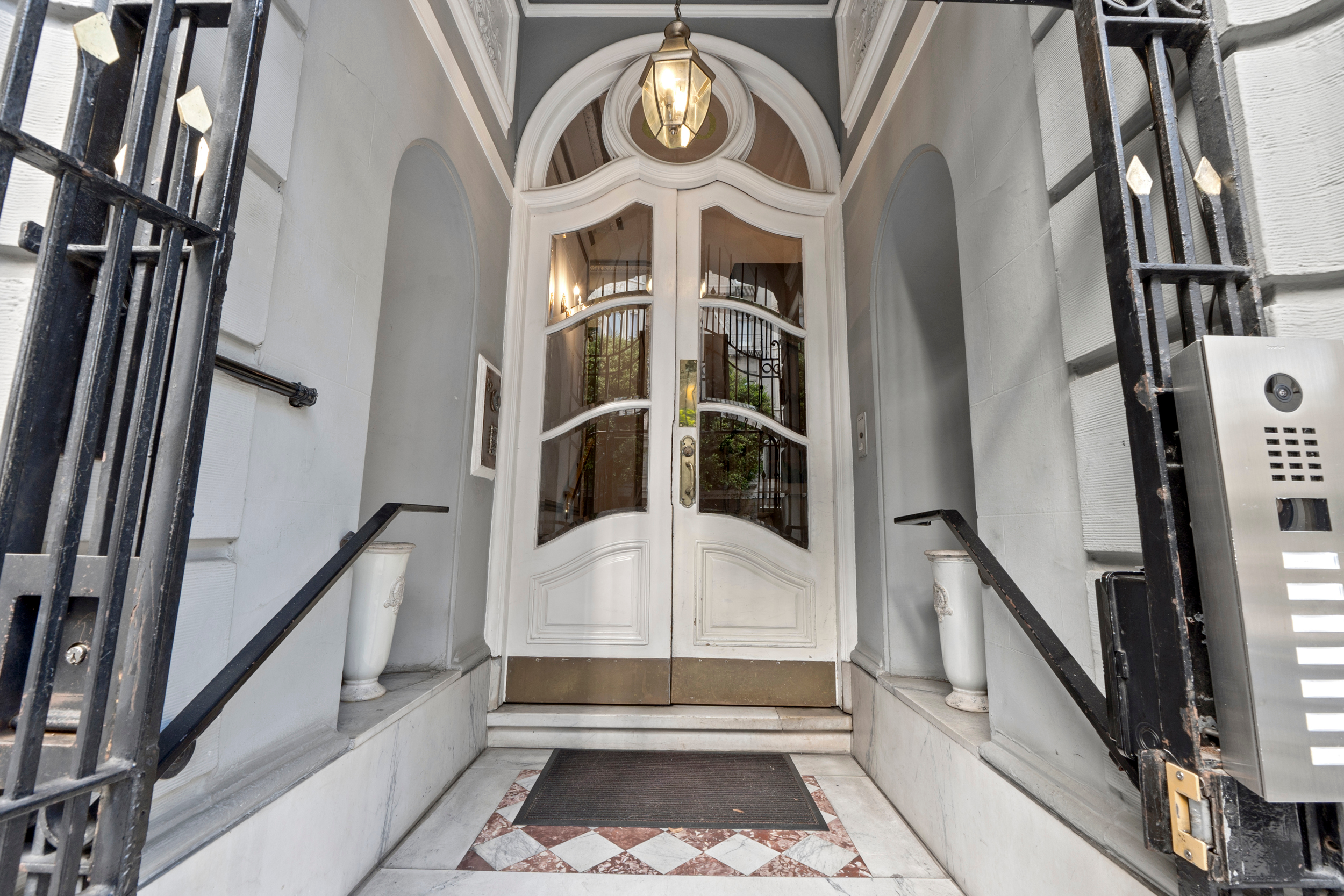 The image shows an elegant entrance with white double doors, a hanging lantern, and steps leading up. Marble tiles and black handrails line each side.