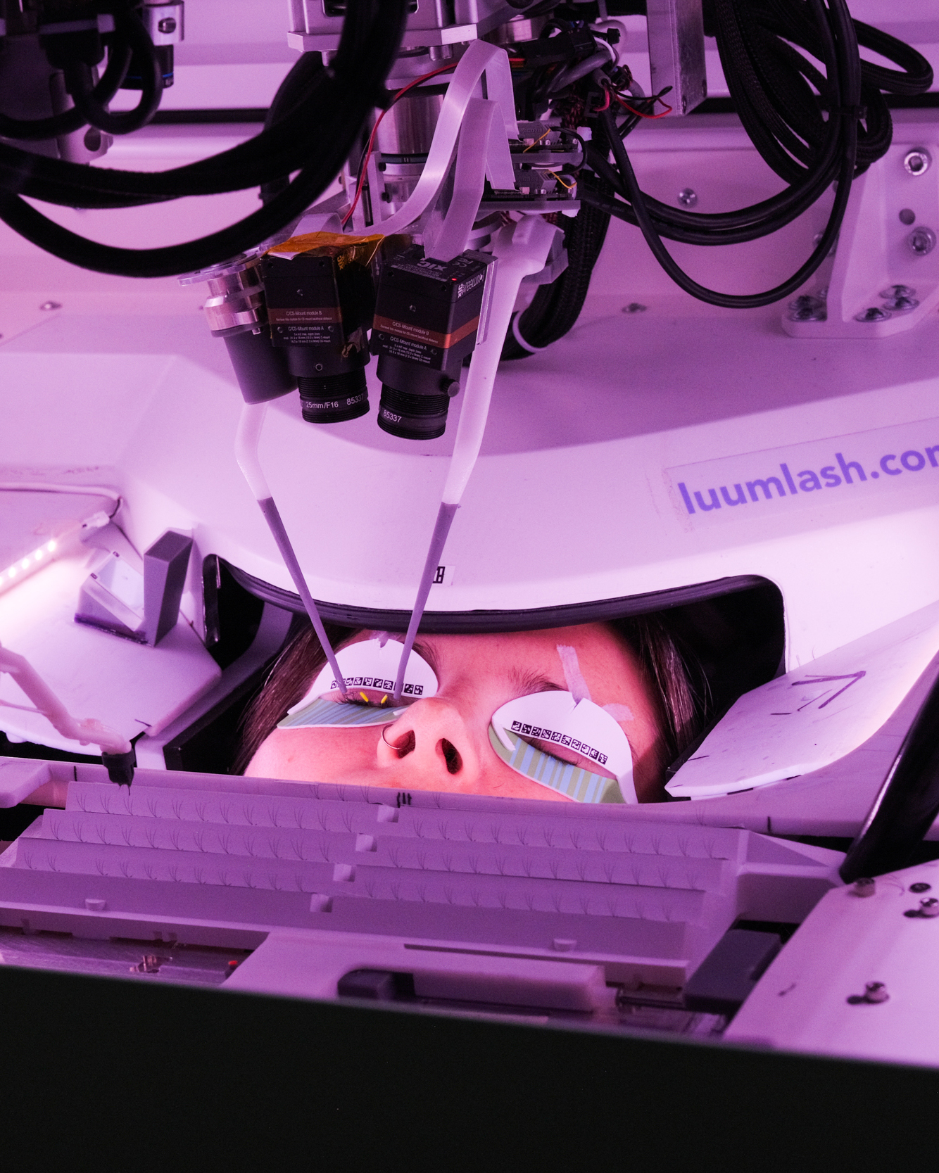 A person is lying under a machine with their head restrained and eyes covered by pads, while two camera-like devices are positioned close to their face, amidst purple lighting.