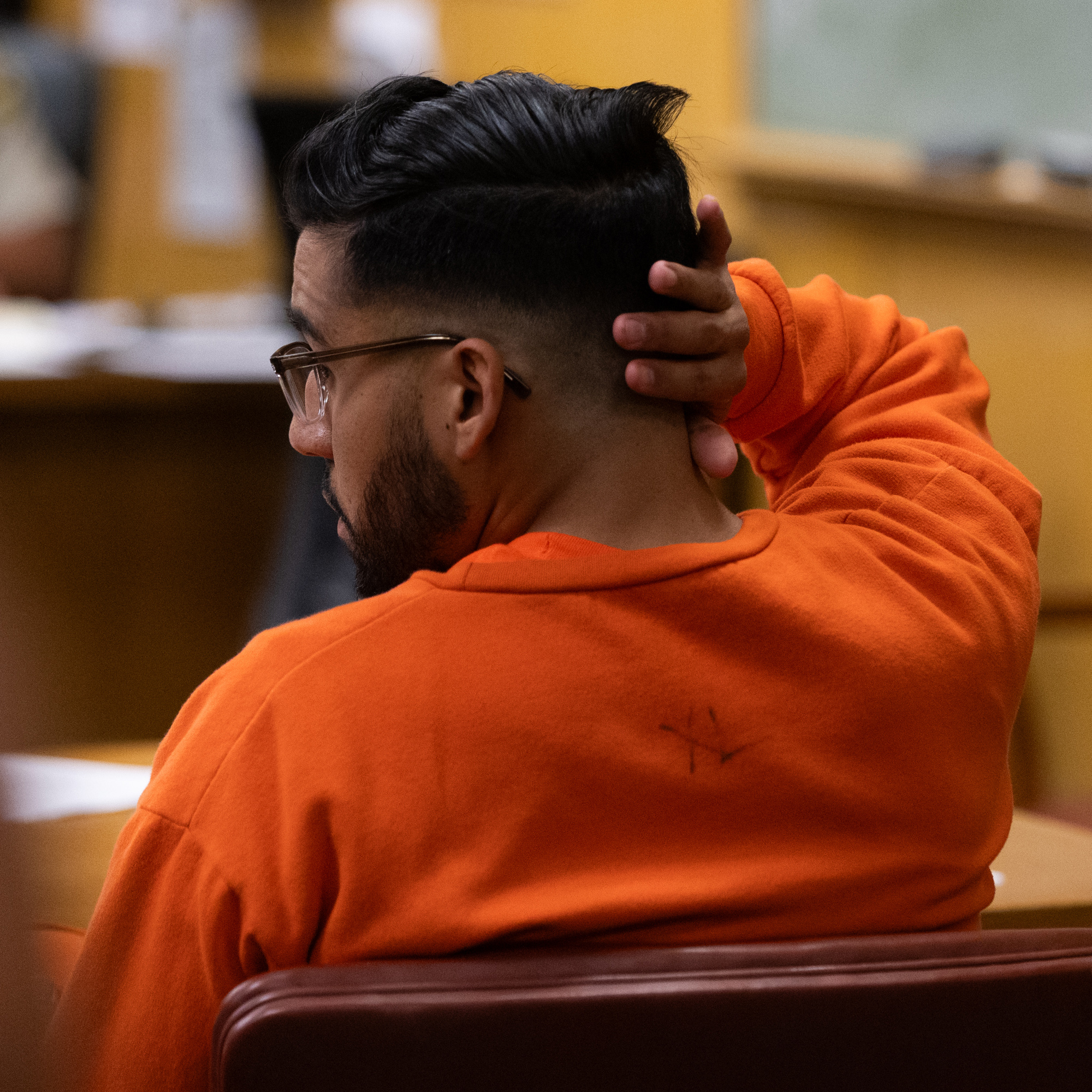 A man in an orange sweatshirt, sits in a courtroom with his hand on the back of his head, and he wears glasses.