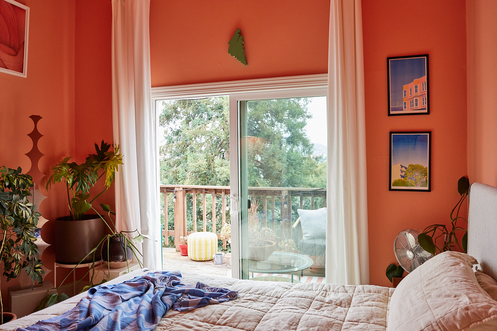 A cozy orange bedroom with plants, artwork, and large sliding doors leading to a deck with a wooden railing and outdoor seating.