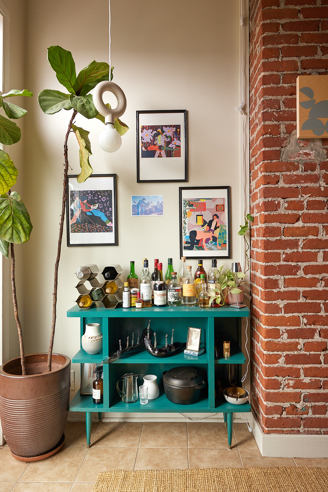 A cozy nook with a teal bar cart stocked with various liquor bottles, a wine rack, and decor. Above, three framed artworks hang on a beige wall beside a potted plant.