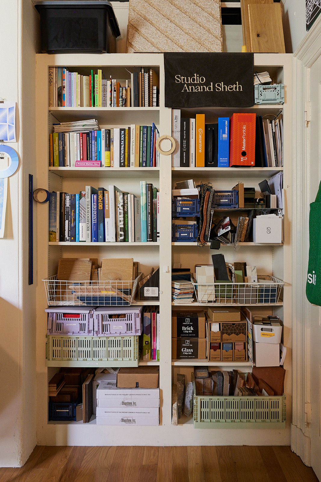 A well-organized shelf holds books, binders, baskets, and assorted materials, including tiles, wood samples, and various project supplies with a banner labeled &quot;Studio Anand Sheth.&quot;