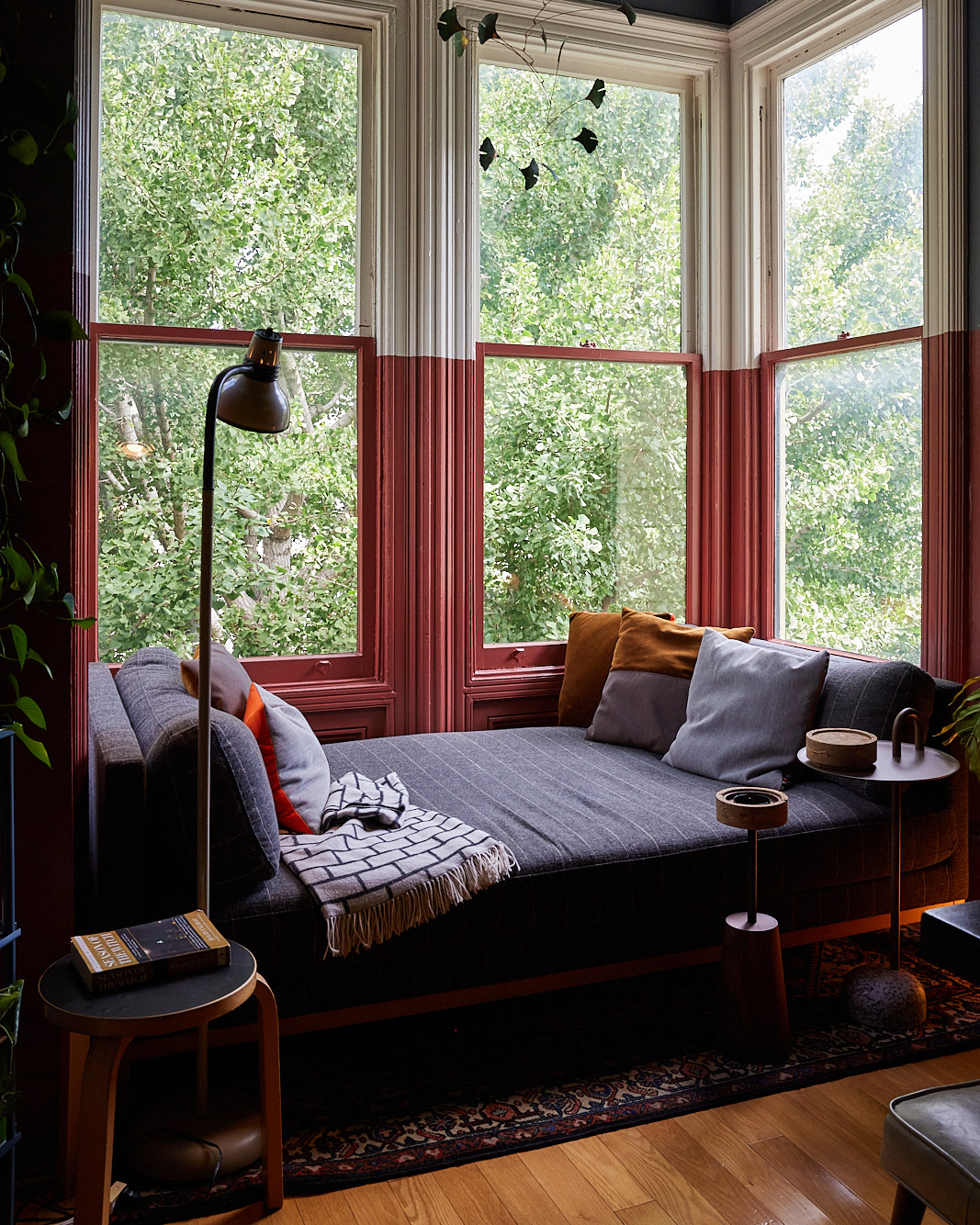 A cozy reading nook with a gray daybed adorned with cushions, set against large windows framing lush green trees. There are side tables with a lamp and a book.