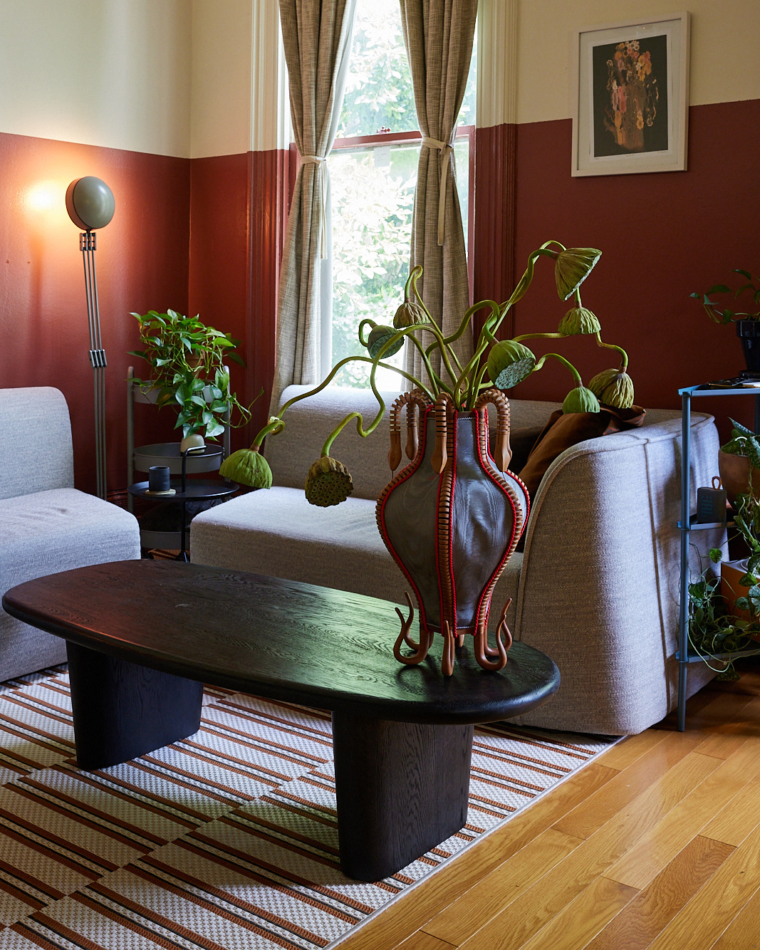 A cozy living room features a gray sofa, a modern black coffee table with a unique vase, a floor lamp, plants, and a window with beige curtains against a maroon wall.