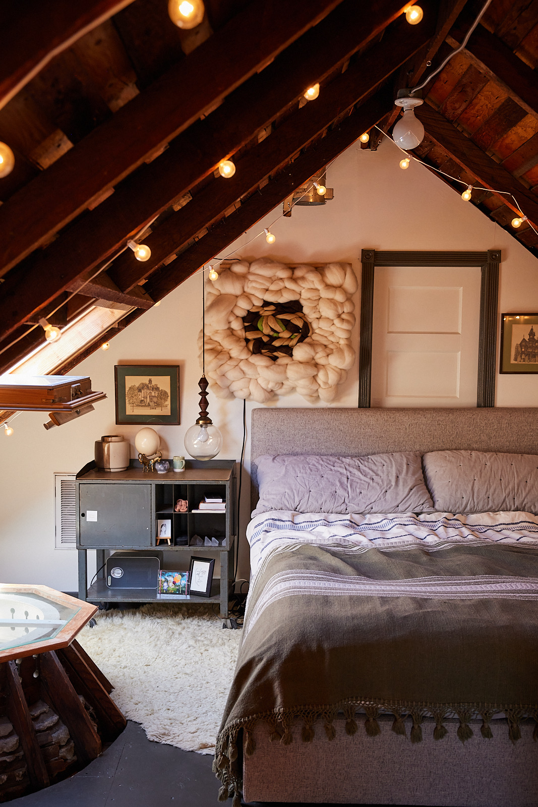 This cozy attic bedroom features exposed wooden beams with hanging string lights, a gray bed, a bedside table with a lamp, decorative wall art, and a fluffy rug.