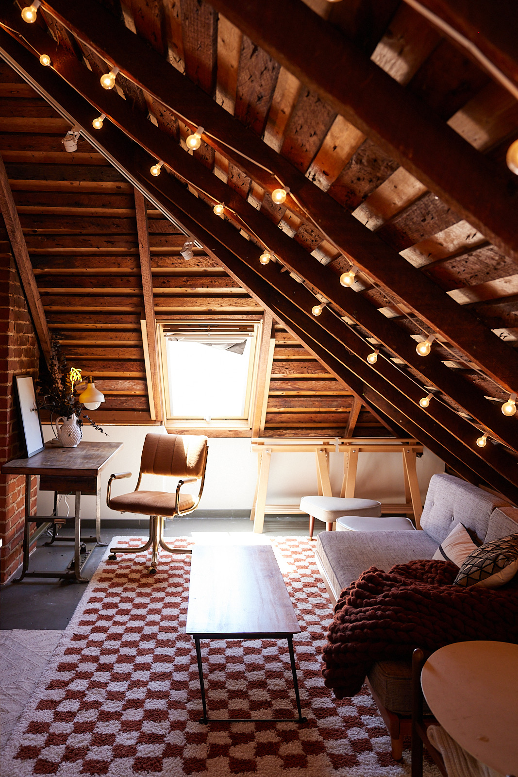 A cozy attic room with exposed wooden beams, string lights, a window, a brown chair at a desk, a gray sofa, and a checkered rug in shades of brown and white.