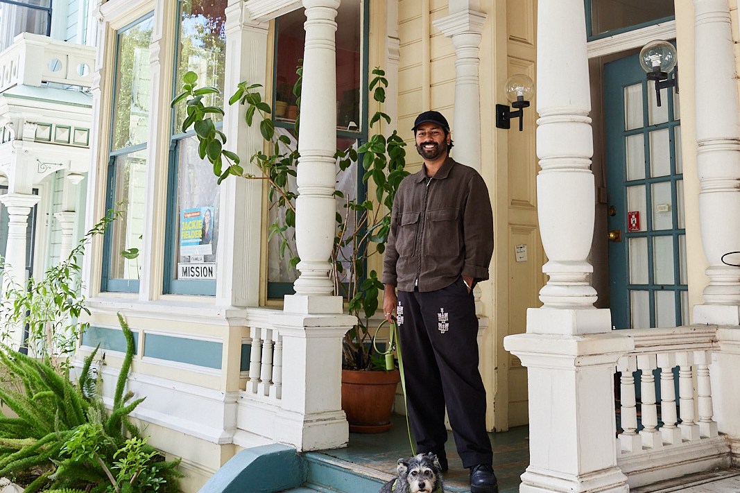 A man and a small dog stand on the steps of a charming Victorian-style house with columns and lush greenery. The man smiles while holding the dog's leash.