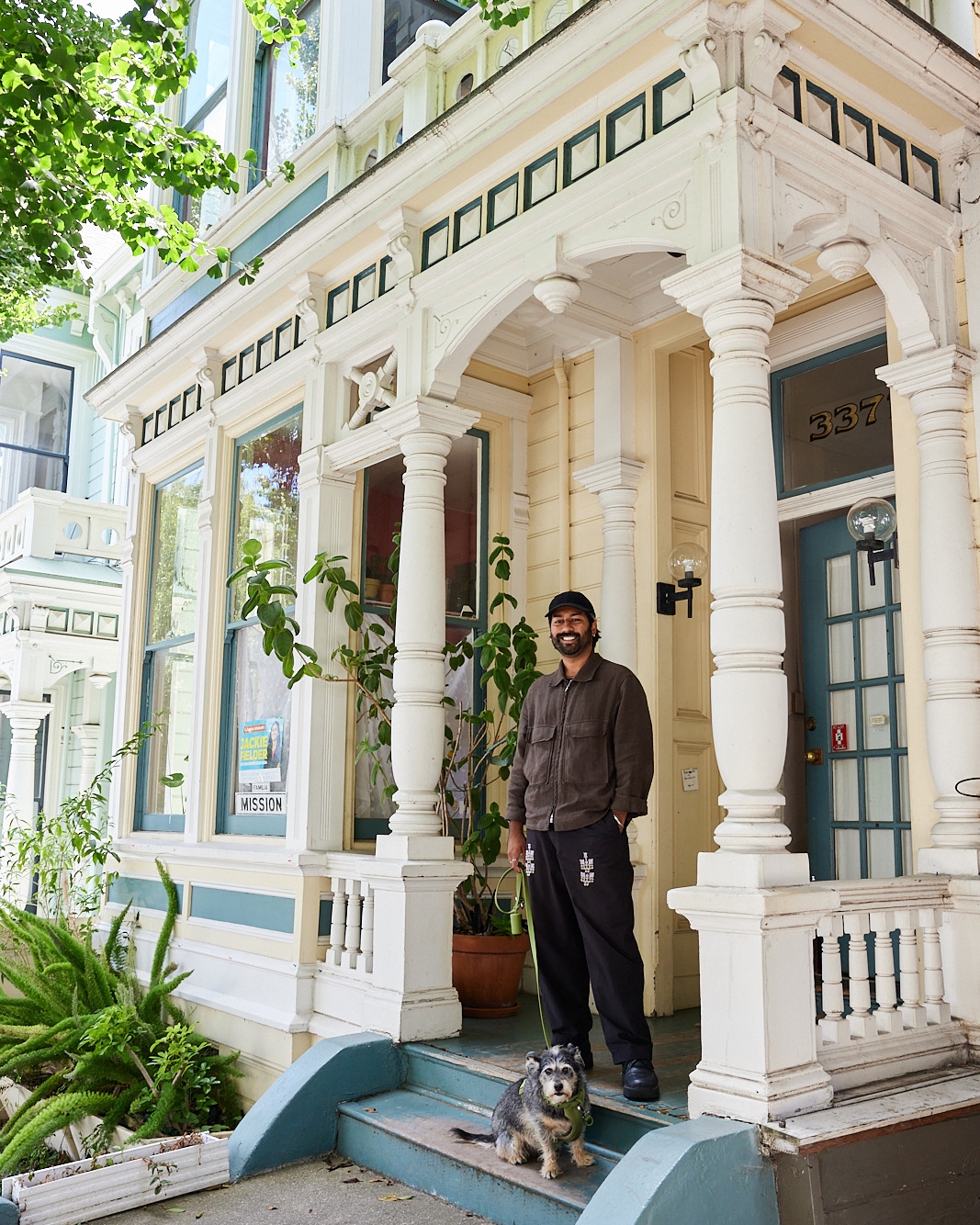 A man and a small dog stand on the steps of a charming Victorian-style house with columns and lush greenery. The man smiles while holding the dog's leash.