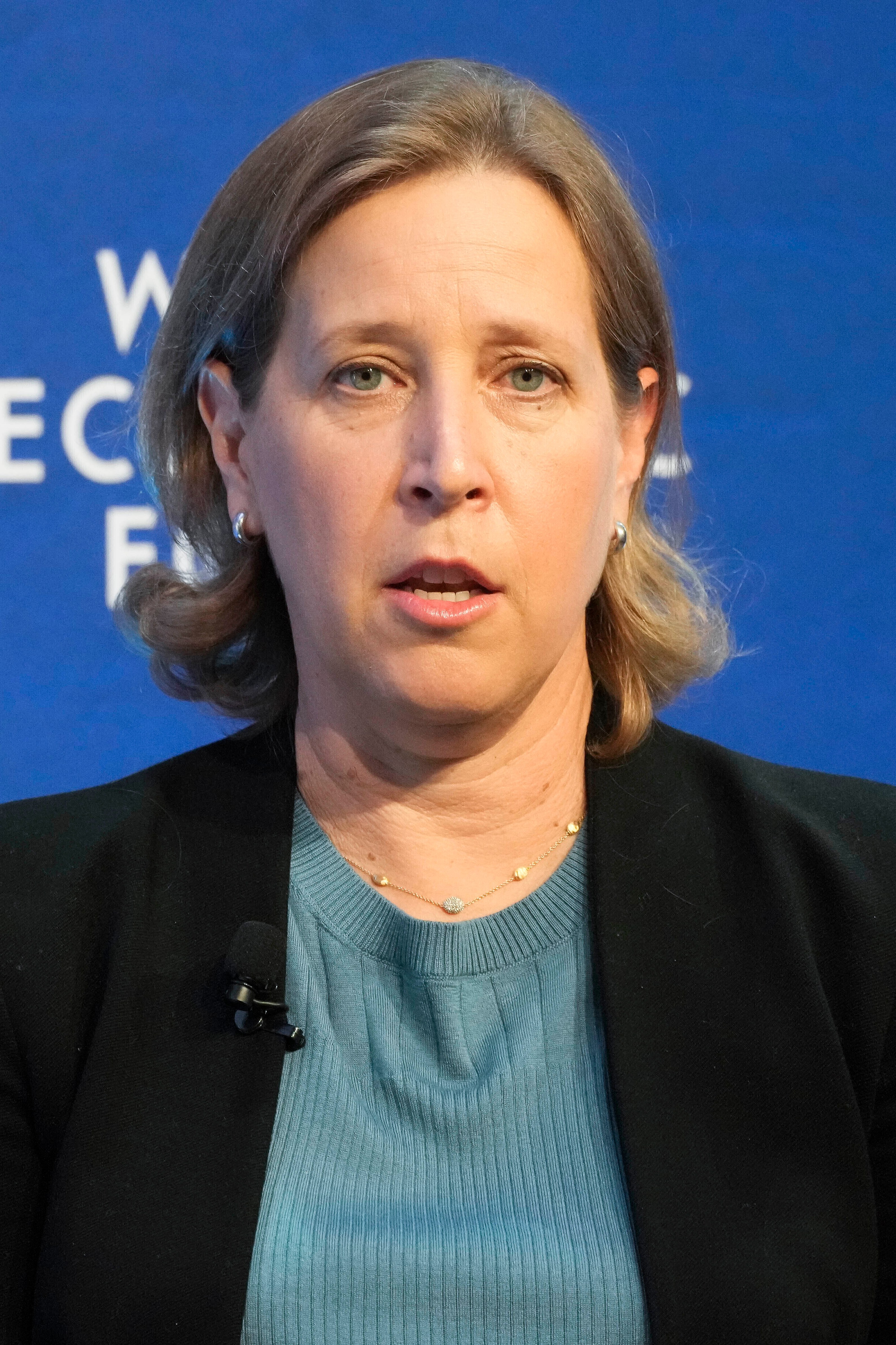 A woman with light brown hair speaks, raising one hand slightly. She is wearing a black blazer over a blue top. The background displays "World Economic Forum."