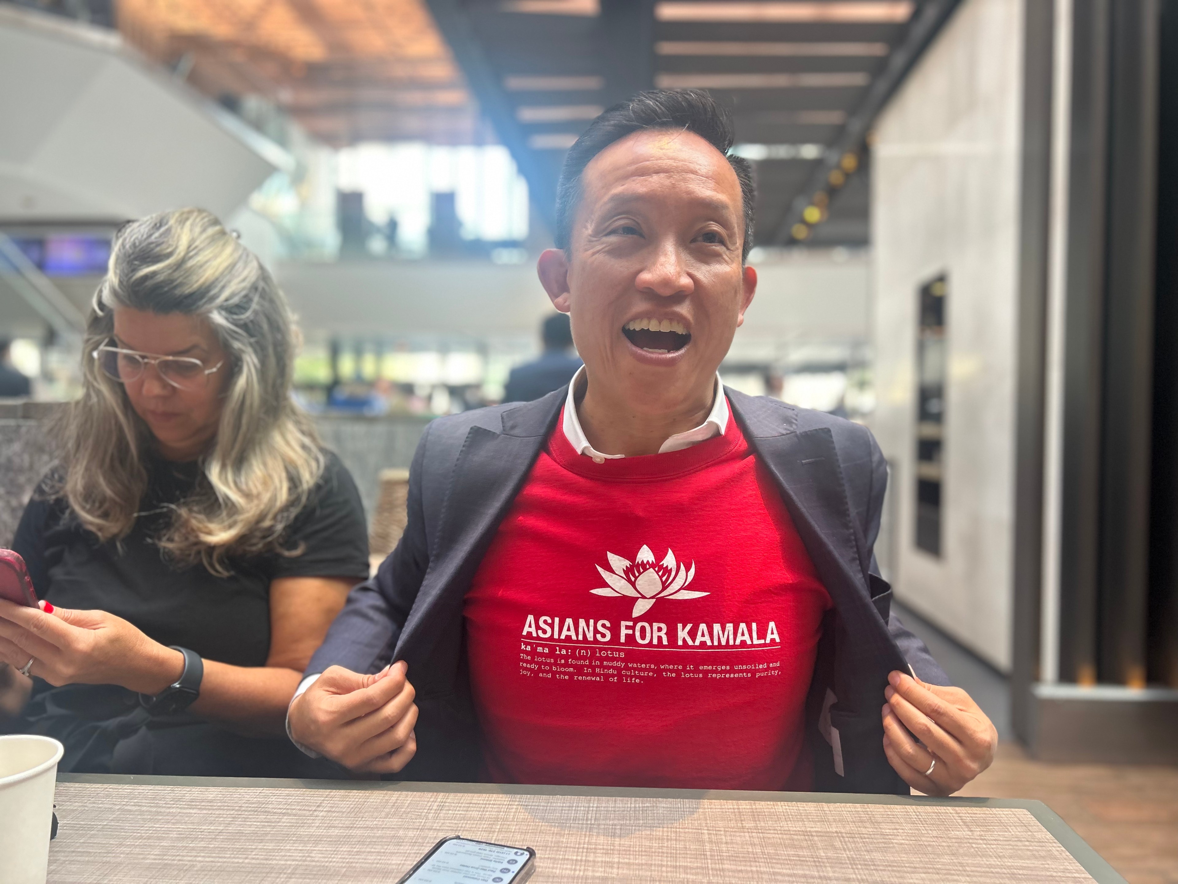 A smiling man opens his jacket to reveal a red shirt reading &quot;Asians for Kamala&quot; with a lotus logo. Beside him, a woman with glasses focuses on her phone.