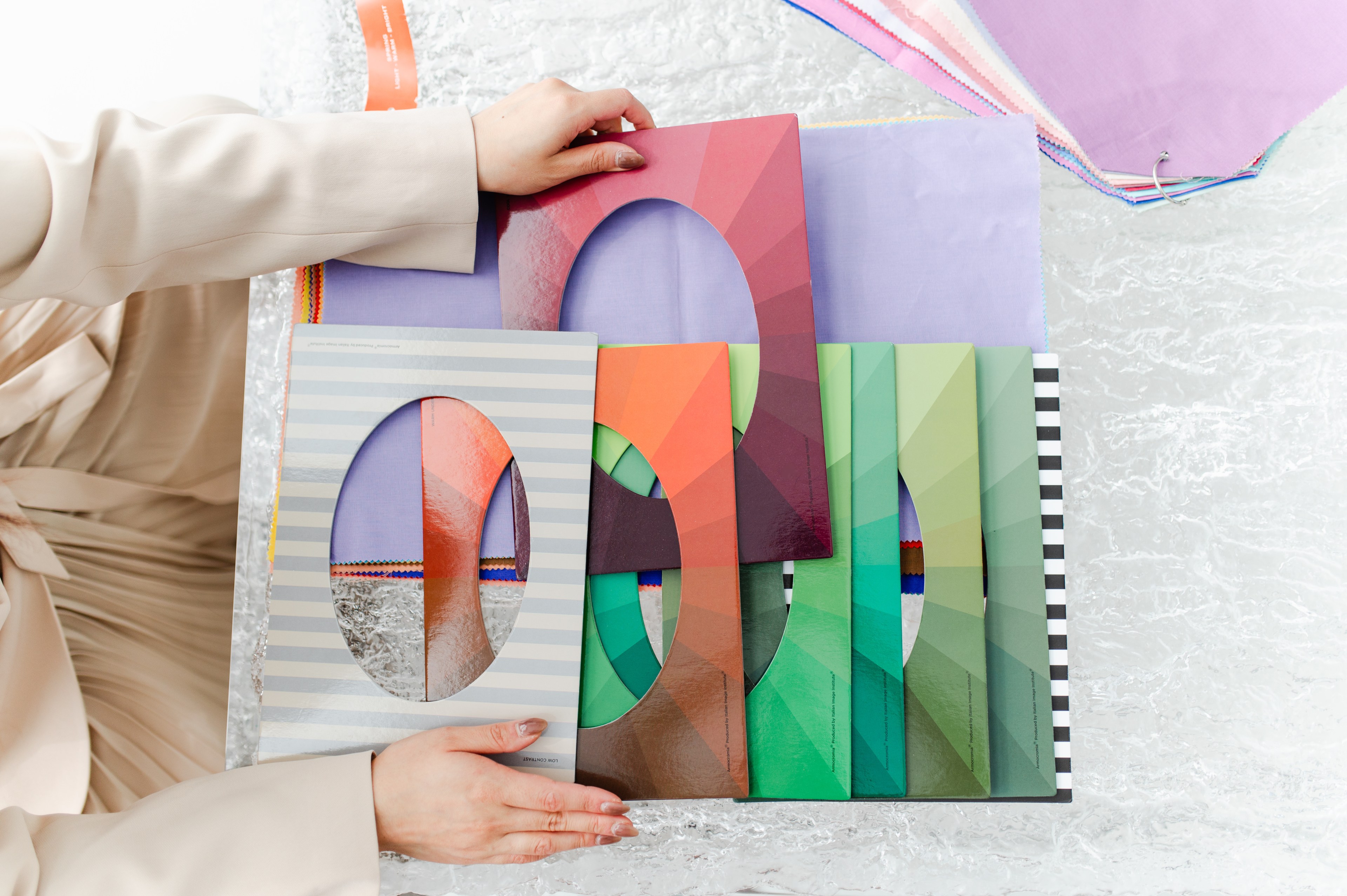 A person in a beige outfit is holding several colorful cardboard frames with oval cutouts, overlaying different shades of fabric samples on a textured surface.