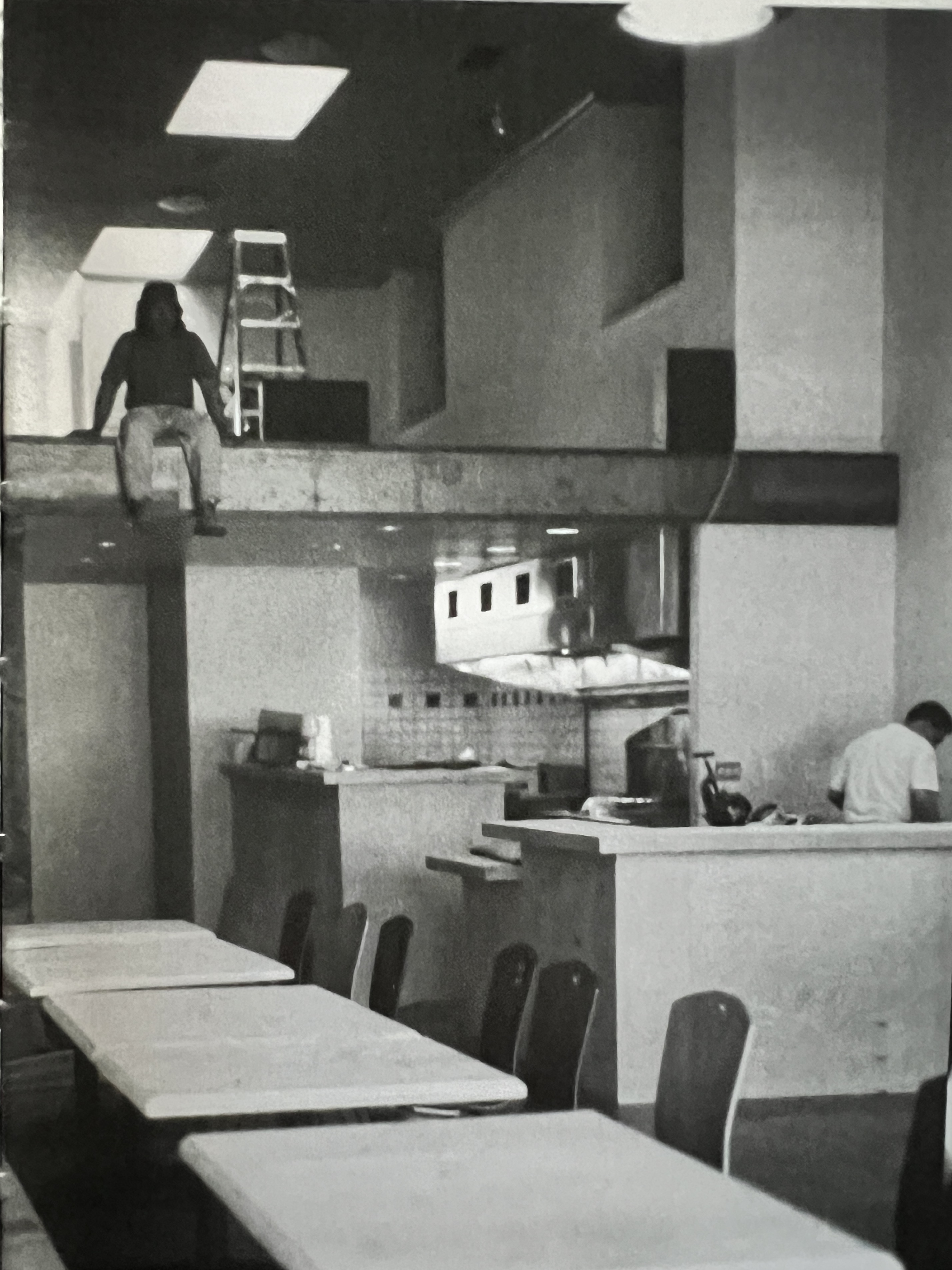 A black and white photo of the interior of a restaurant, where a man sits with his feet dangling off a loft.