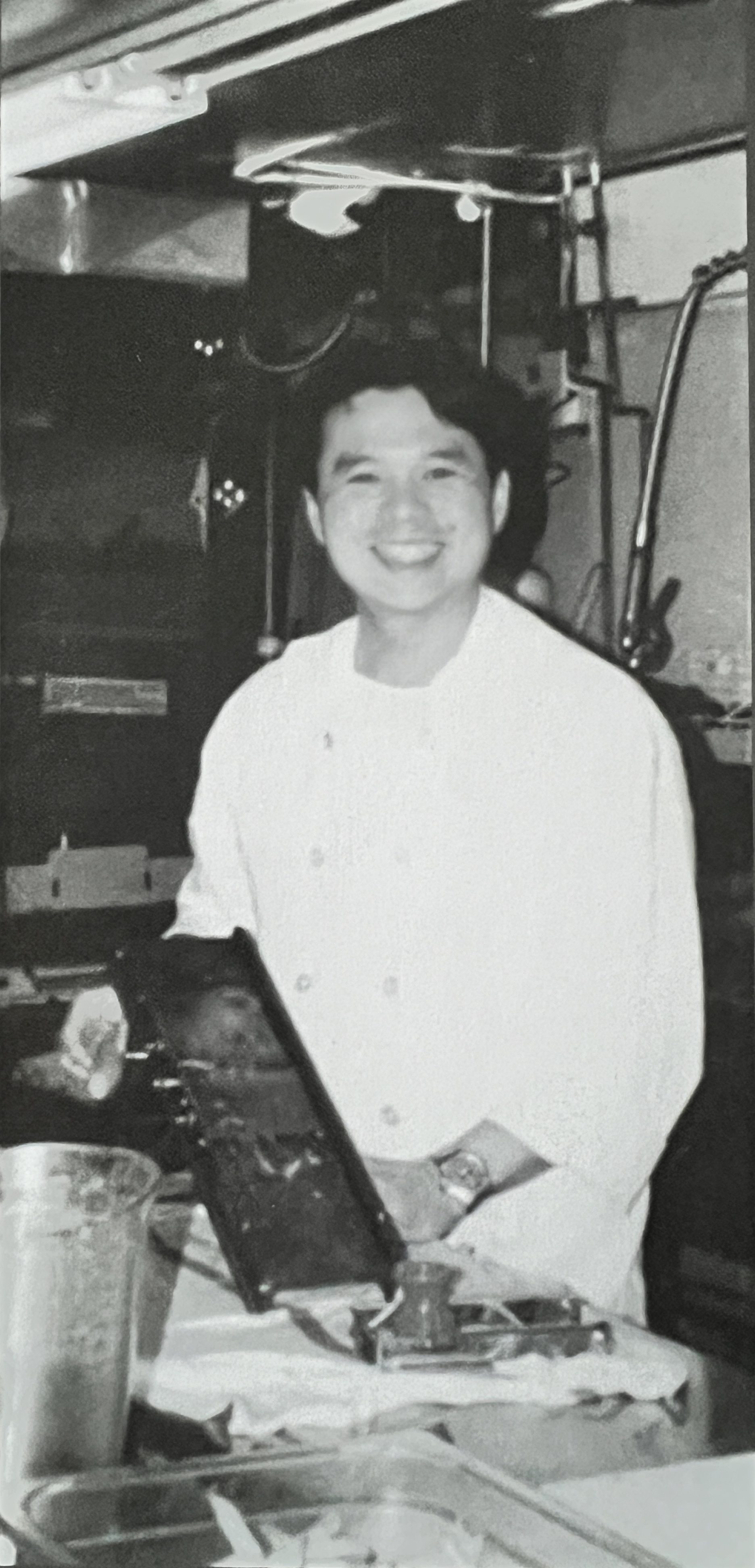 A black and white photo of a young man wearing a white chef coat as he smiles while standing in a kitchen.