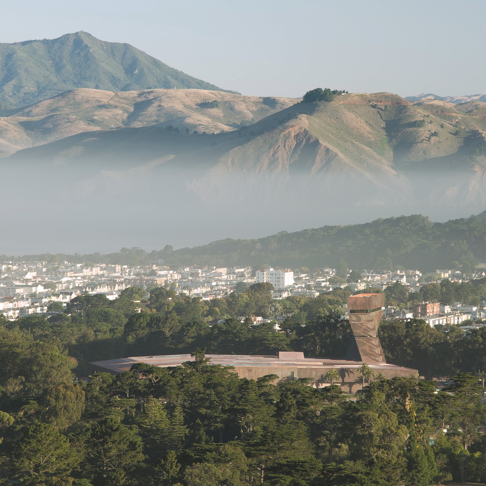 A copper colored museum sits within a green canopy in the foreground. In the background, lush mountaintops poke out of the fog. 