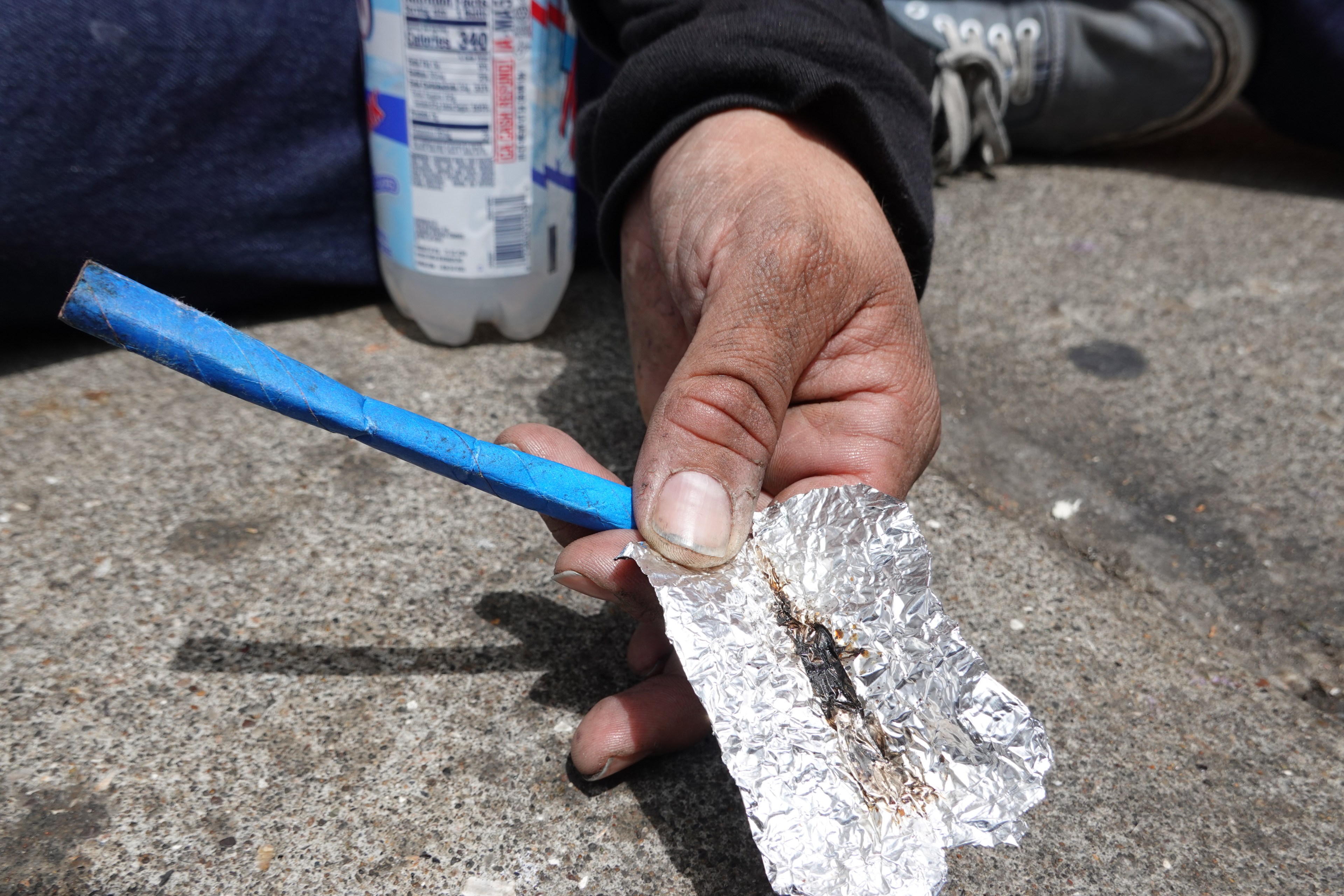 A person's hand is holding a crumpled piece of foil and a worn blue straw, with a bottle in the background and seated legs on a concrete surface.