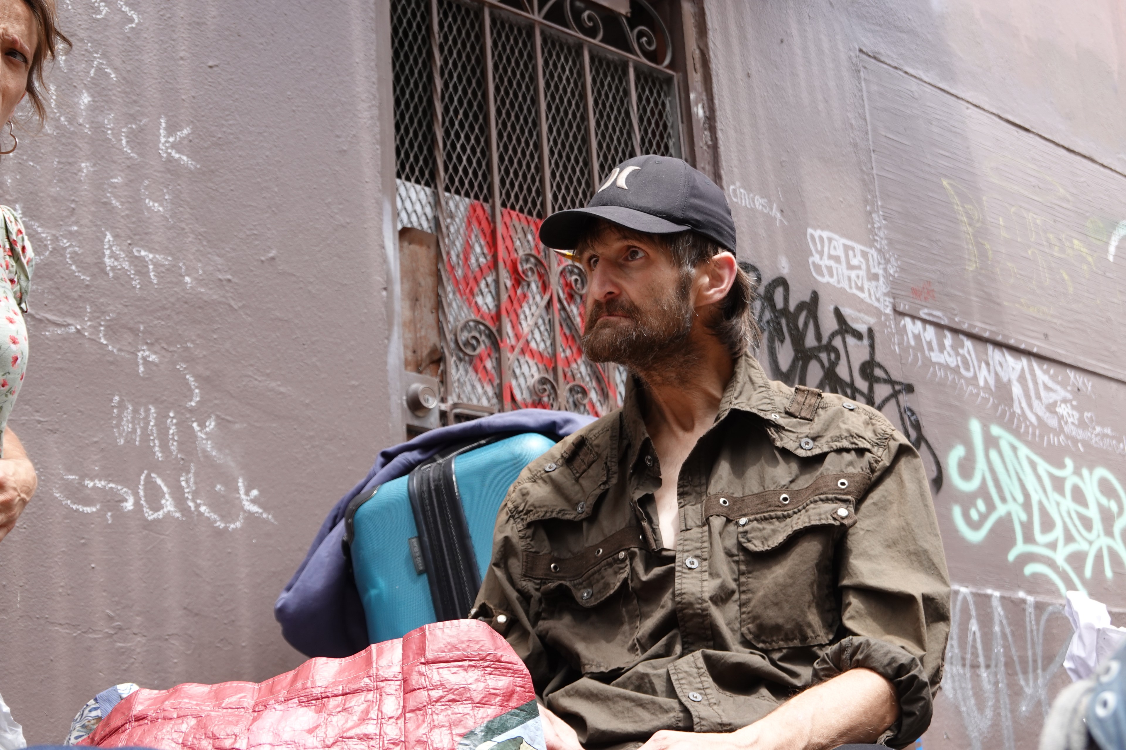 A man in a worn-out shirt and cap sits by a wall covered in graffiti with a suitcase and blanket in front of him. A woman partially appears on the left.