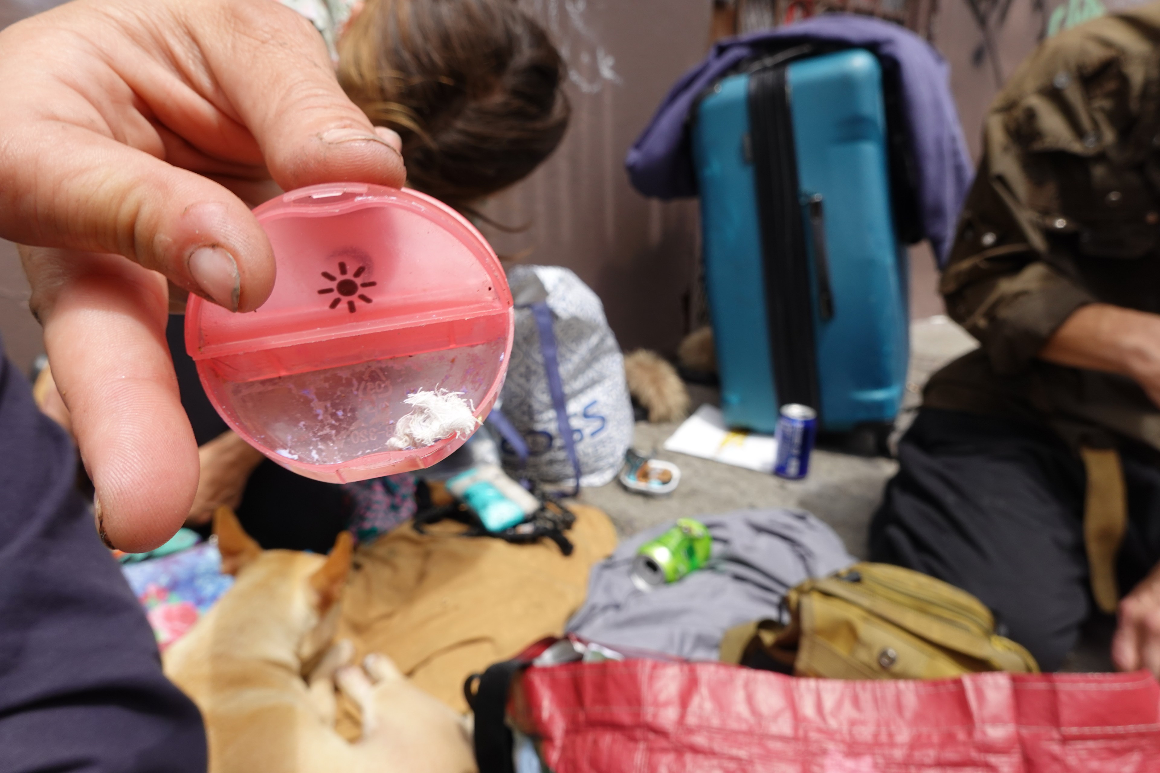 A person holds a small pink container with white residue. In the background, people sit near a suitcase, scattered bags, a dog, and drinks.