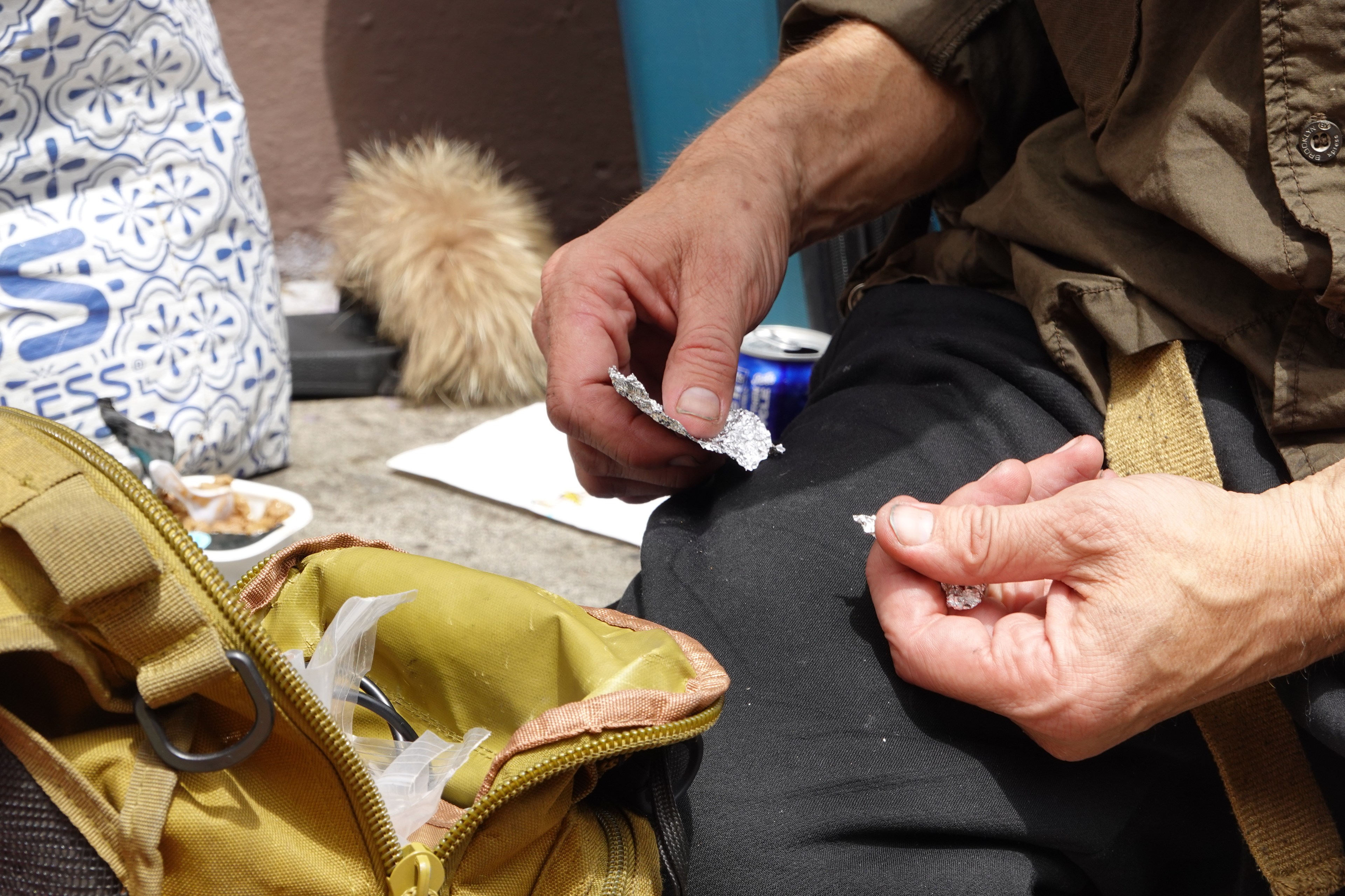 A person is sitting on the ground with a yellow bag open beside them. They're holding small pieces of crumpled aluminum foil. Nearby, there's a can of soda and a patterned bag.