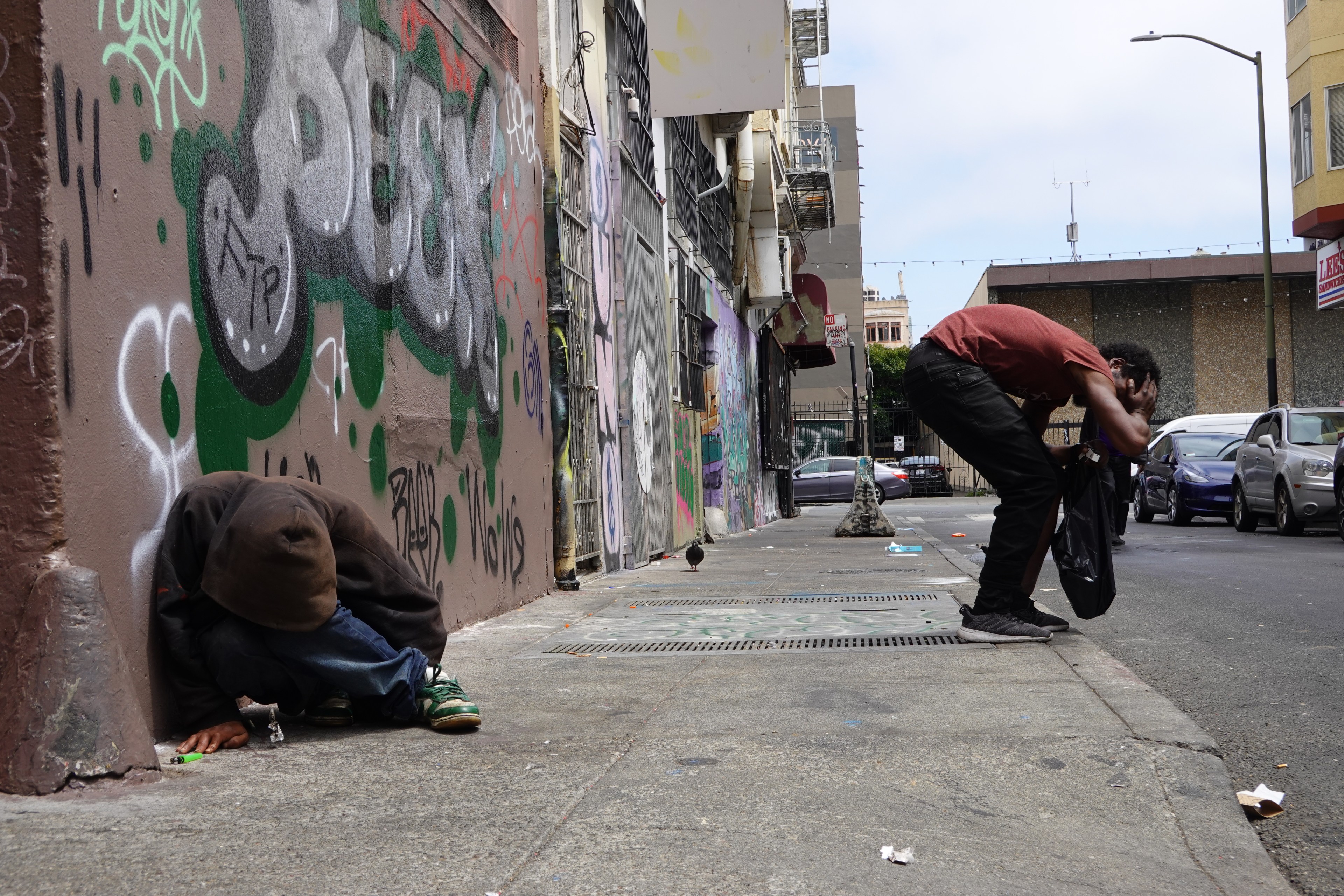 Two individuals in an urban alley with graffiti; one is sitting against a wall, and the other is bent over, appearing distressed. Two parked cars are in the background.