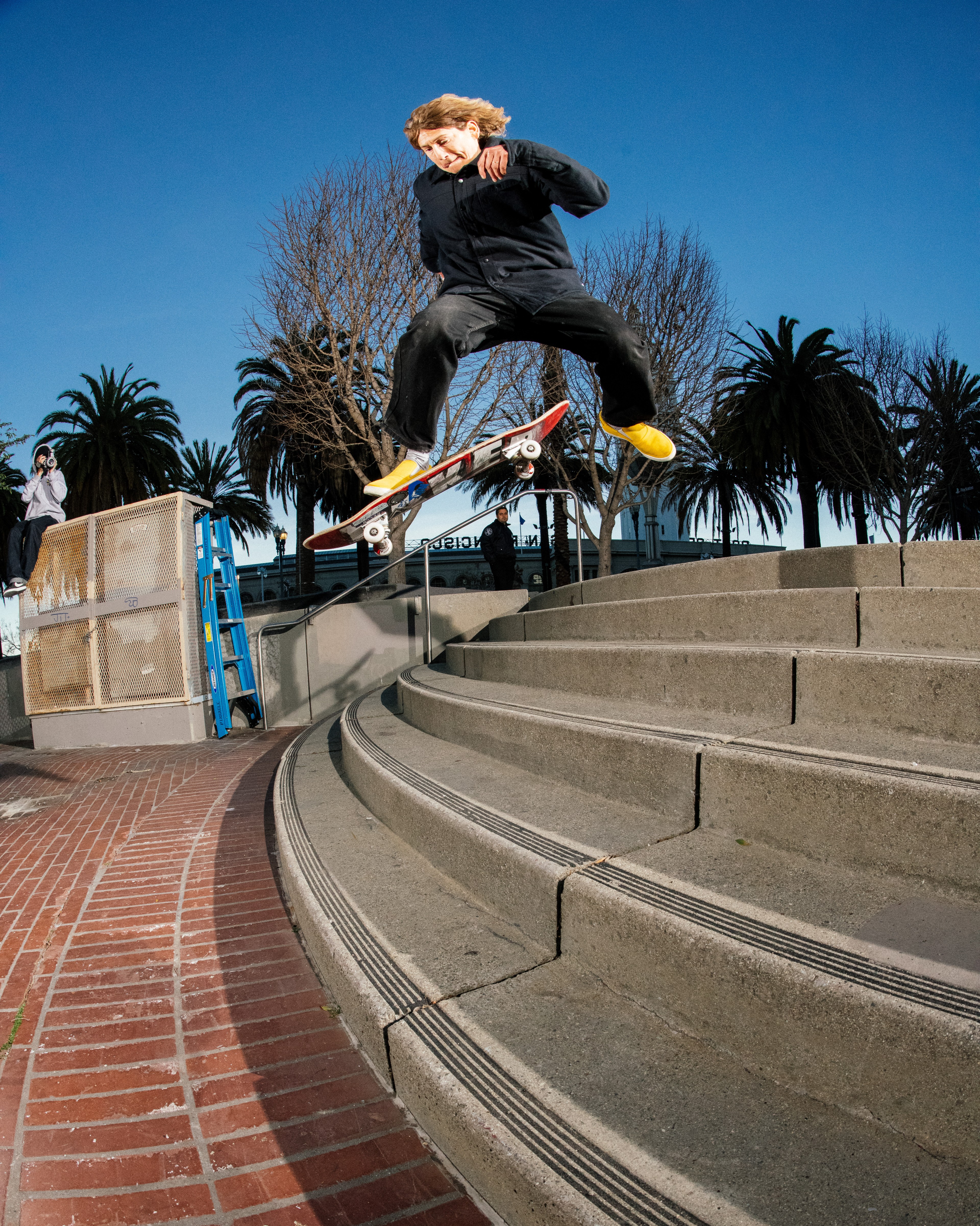 A person skateboards mid-air down concrete steps under a sunny blue sky, surrounded by palm trees and bare trees, while another person films in the background.