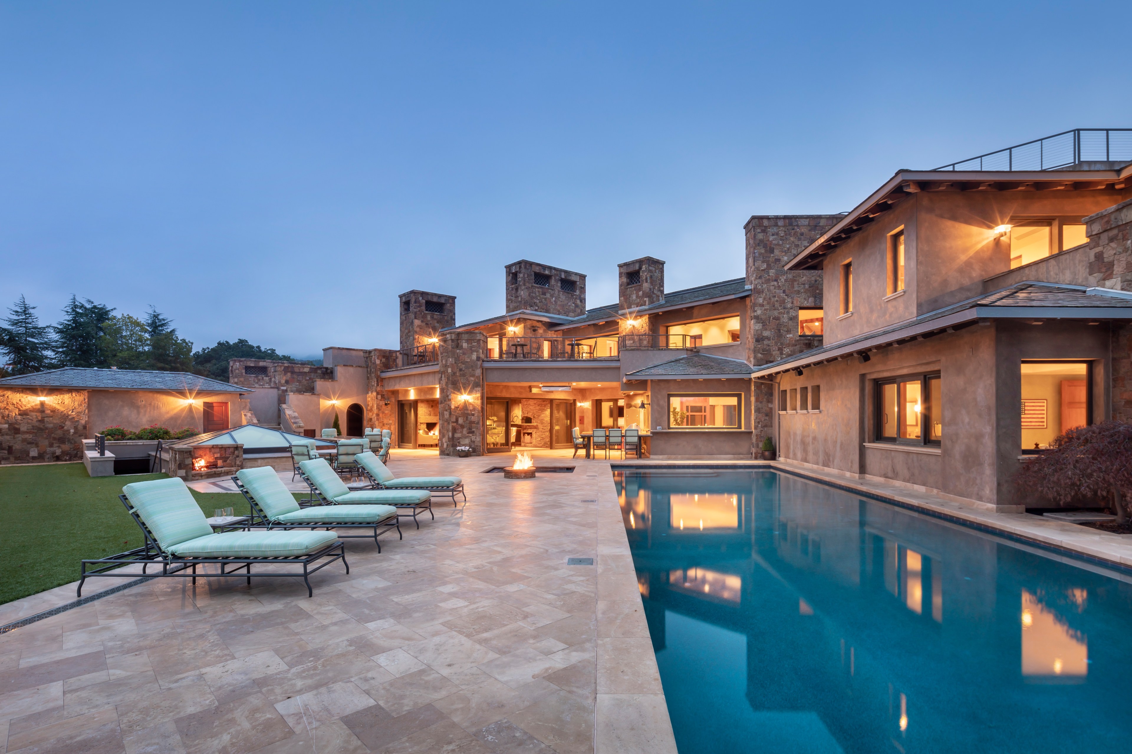 A luxurious, modern house with a large swimming pool, sun loungers, and a patio area, illuminated by warm outdoor lighting at dusk. The building has a stone facade and multiple windows.